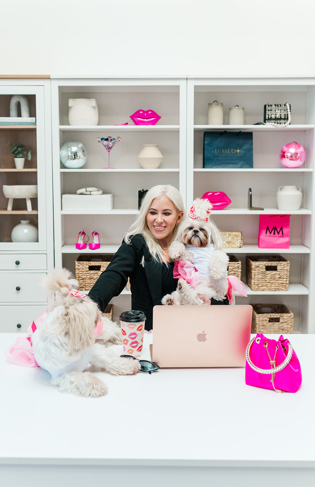 Woman with two small dogs at a white desk, laptop, pink bag, and colorful decor in background.