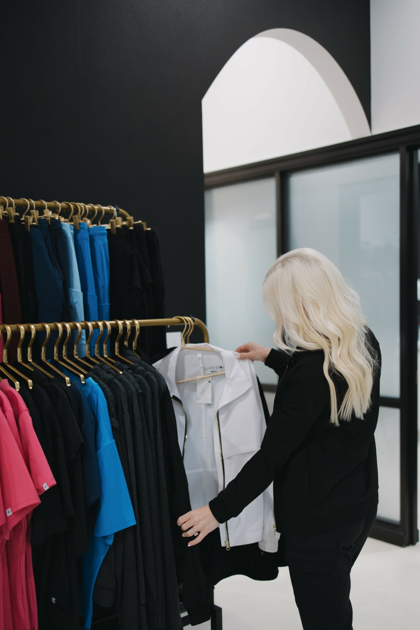 Woman with long blonde hair shopping for clothes in a store, browsing through shirts on a rack, with large windows behind her.