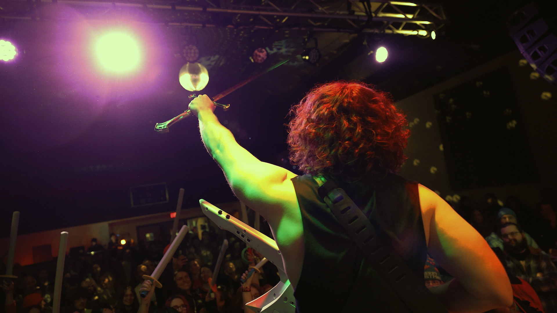 Performer with curly hair on stage, holding a microphone, with bright stage lights overhead and audience in background.
