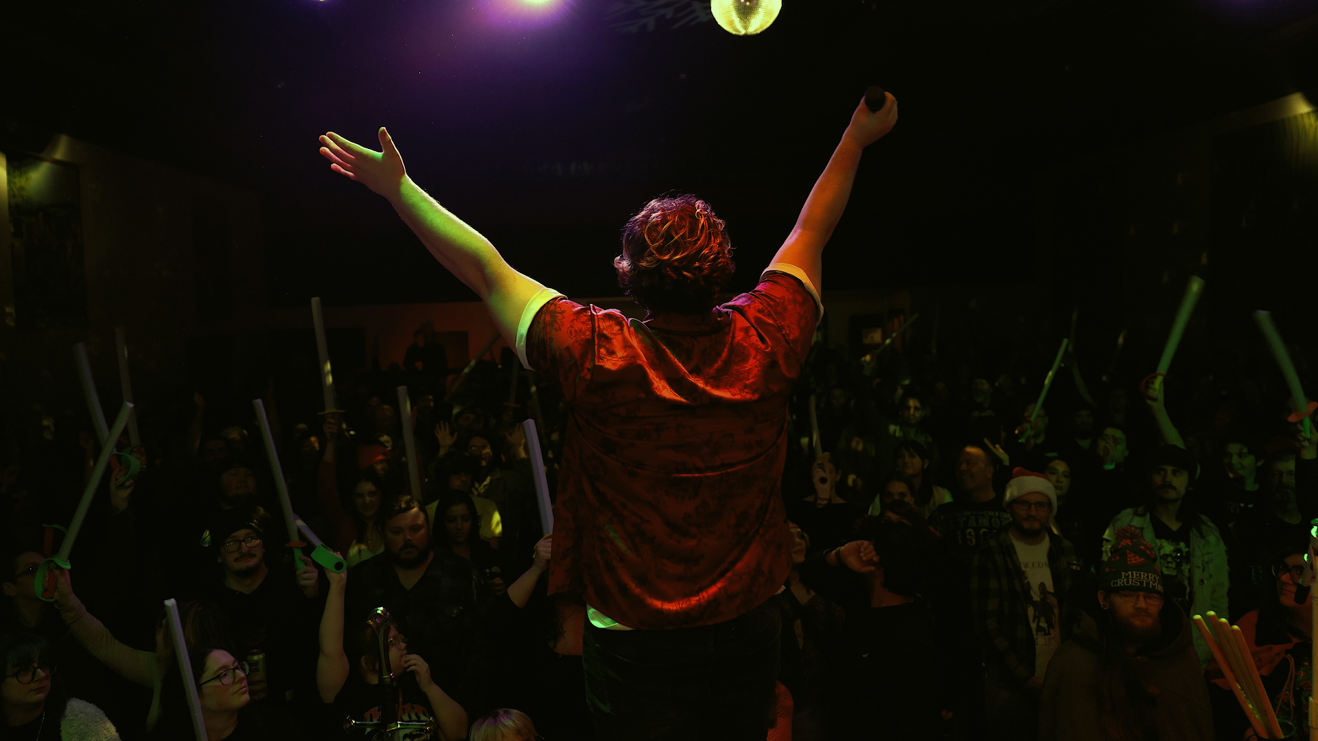 Performer with arms raised on stage facing an audience in a dark venue with lights and glow sticks.