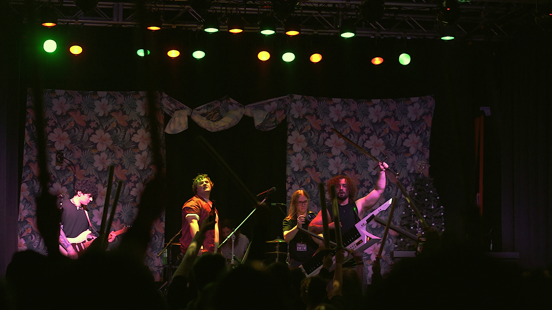 Four performers on stage with floral backdrop, colorful lights overhead, and musical instruments, engaging in a performance.