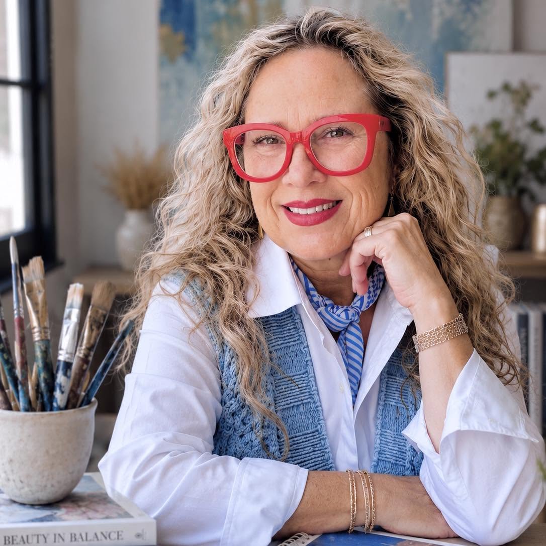 Woman with curly hair and red glasses smiling, sitting at a desk with art supplies, in a bright room.