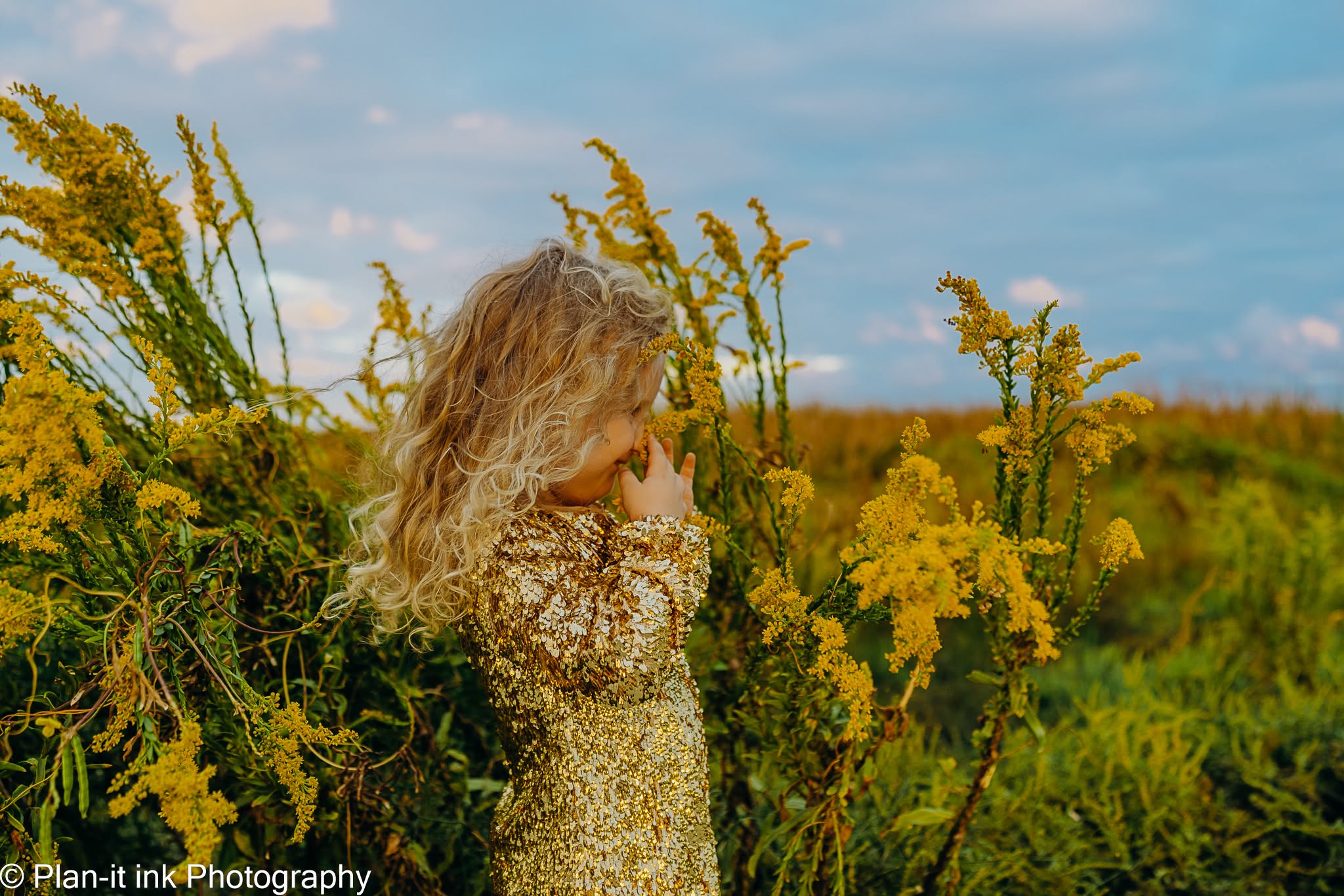 Child with curly hair standing among yellow flowers outdoors, facing away, with a blue sky and green landscape in background.
