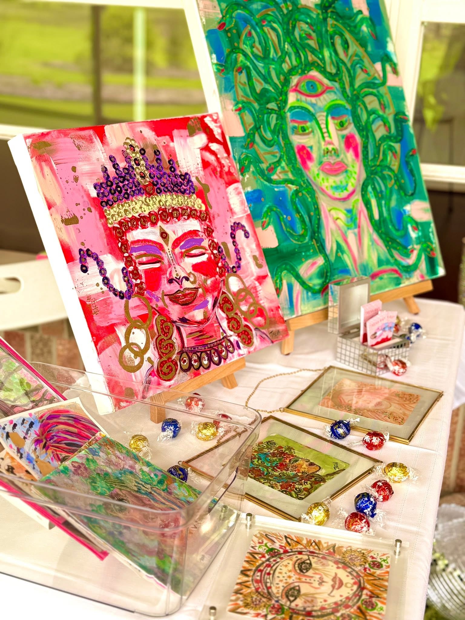 Two colorful paintings of faces with vibrant details, displayed on a table with jewelry and art supplies.