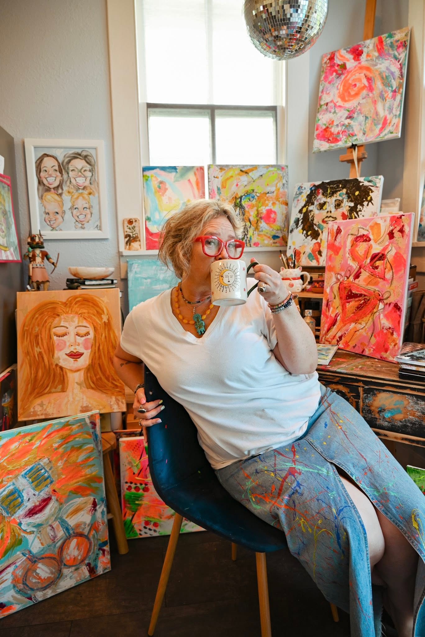 Woman with red glasses sitting in an art studio, holding a mug, surrounded by colorful paintings and artwork.