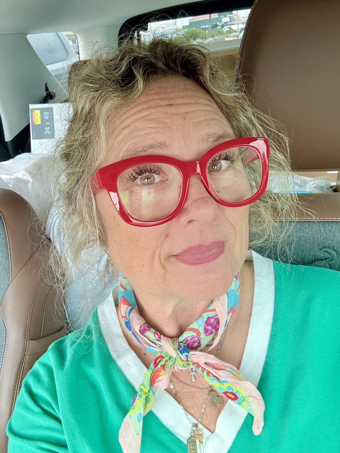 Woman with curly hair wearing red glasses and a colorful scarf, sitting in a car.