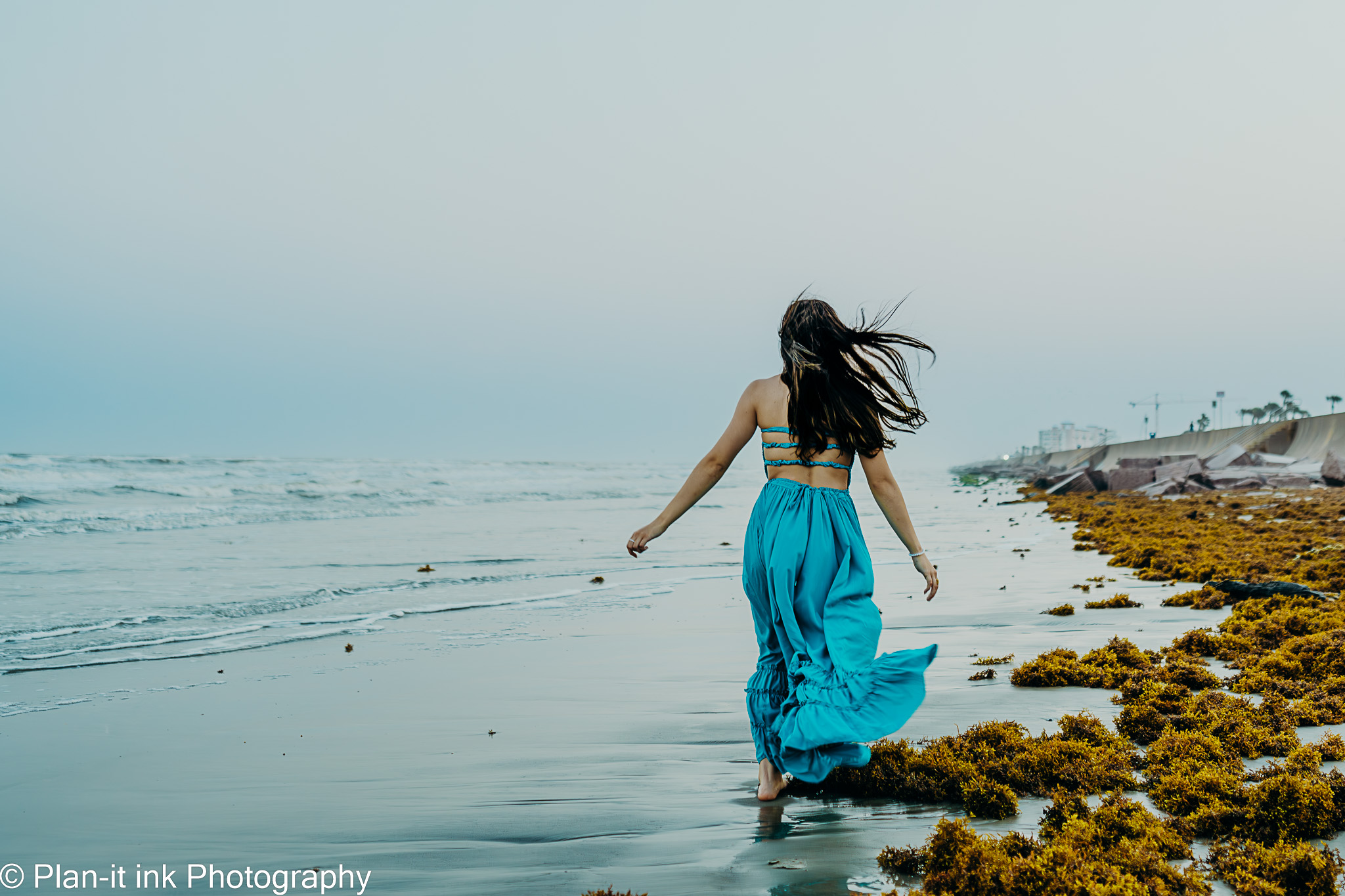 Person walking along the beach with long hair, wearing a blue skirt and top, sea and shoreline in background