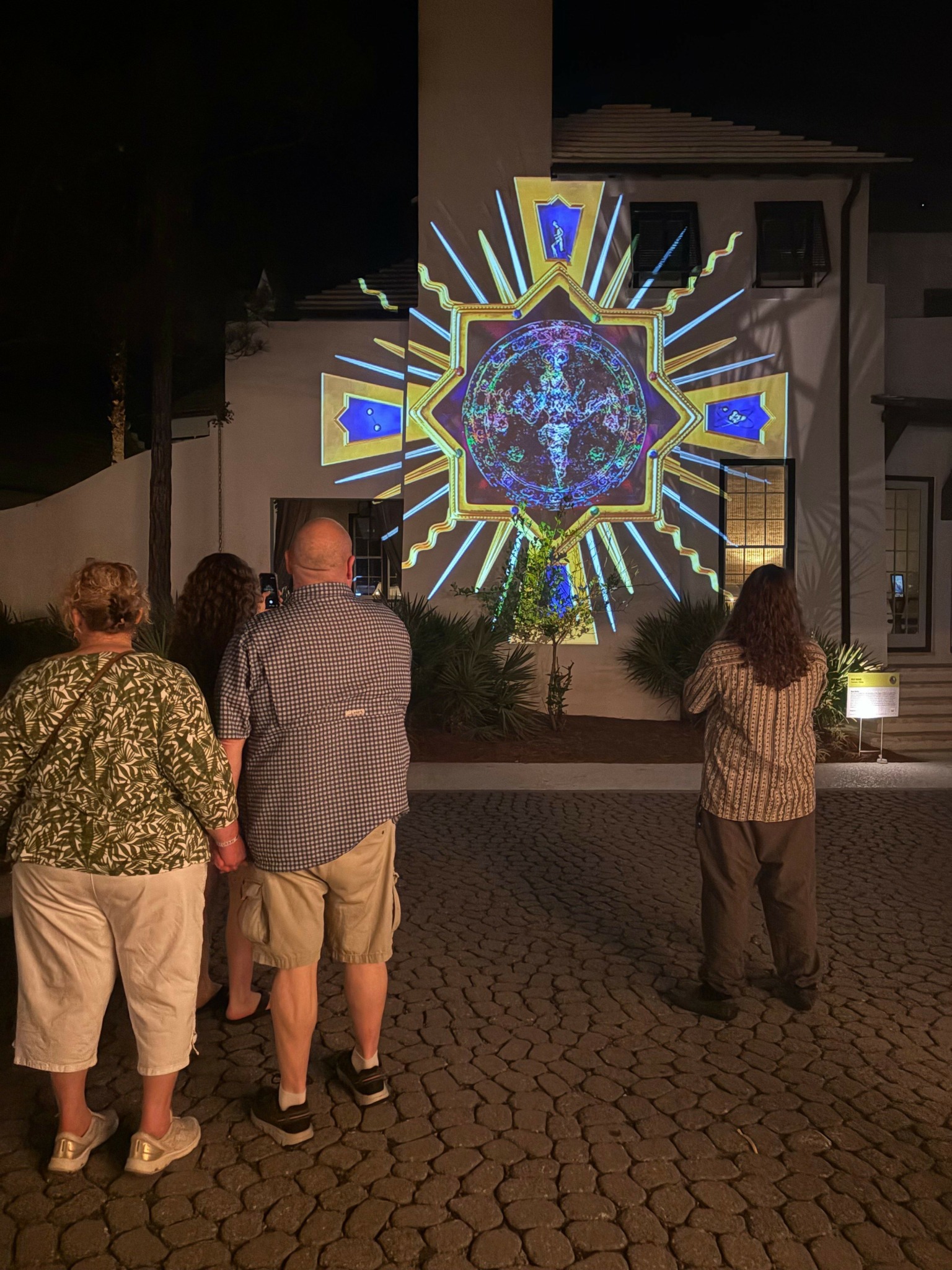 Two people stand in front of a large illuminated star-shaped display on a building wall at night, with a woman in a patterned top and beige pants in the foreground.