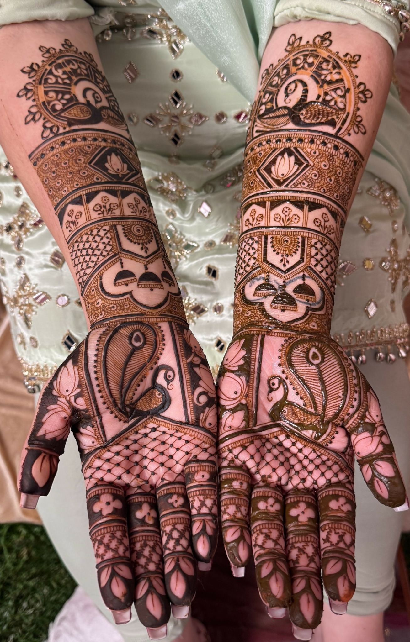 Person with intricate henna designs on both hands and forearms, wearing a light-colored, embroidered outfit, with hands open showing palms.