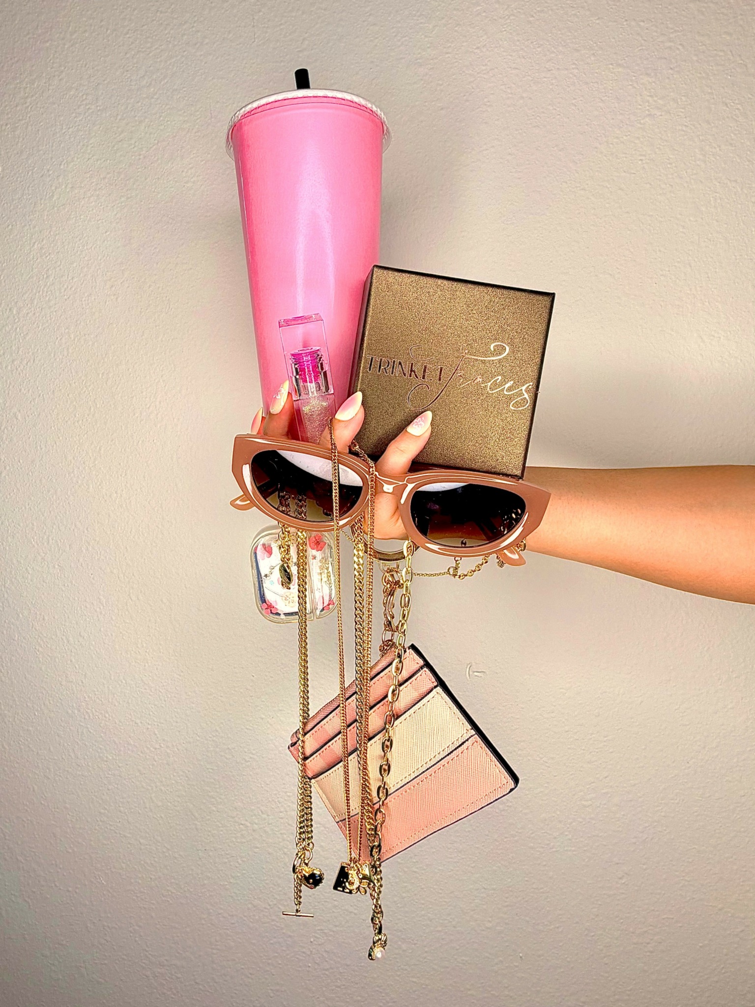 Hand holding a wall-mounted organizer with pink tumbler, sunglasses, jewelry, and small purse, against a plain wall.