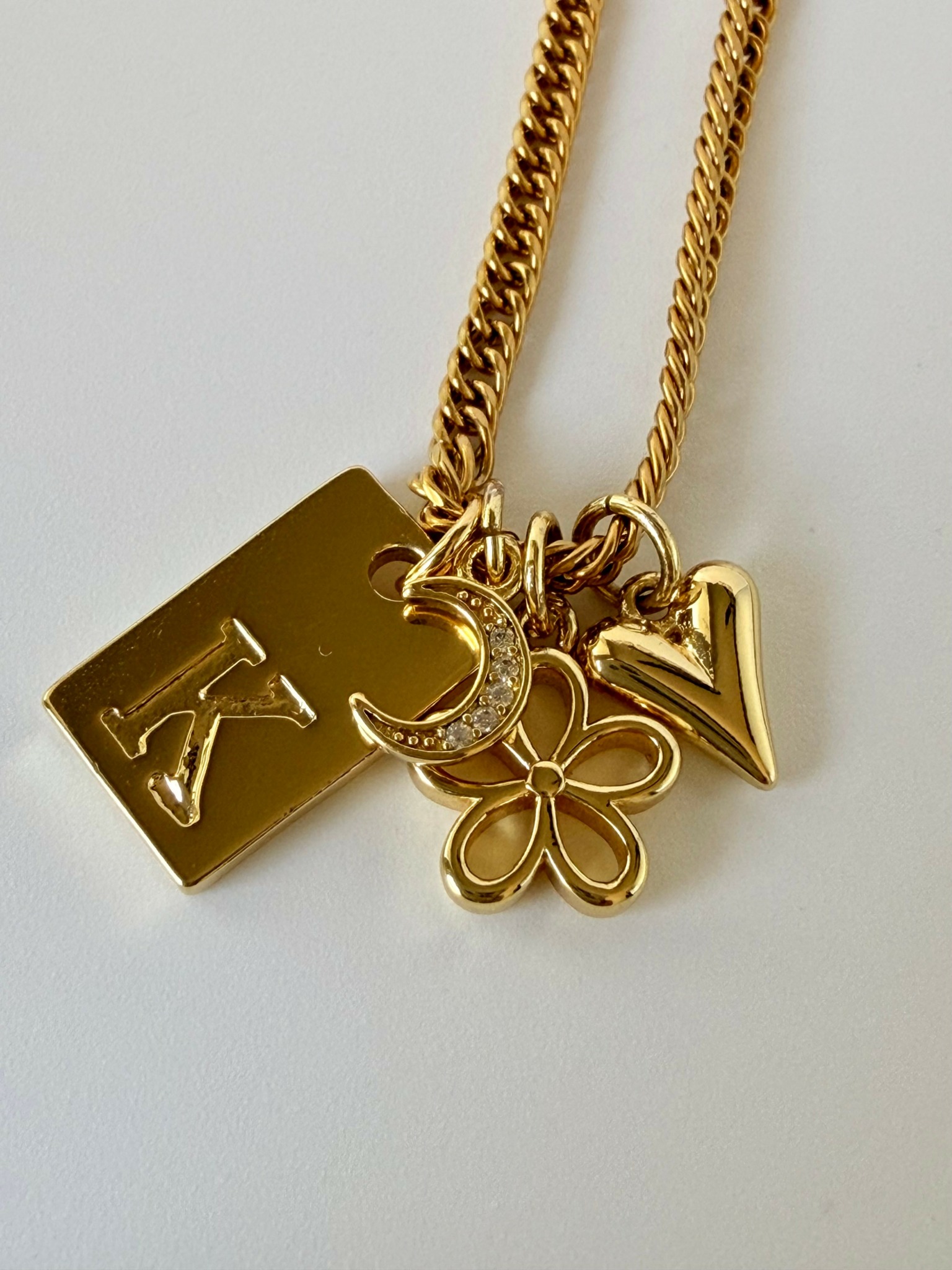 Gold chain with four charms: a rectangular tag with a letter, a crescent moon, a flower, and a heart-shaped letter V.