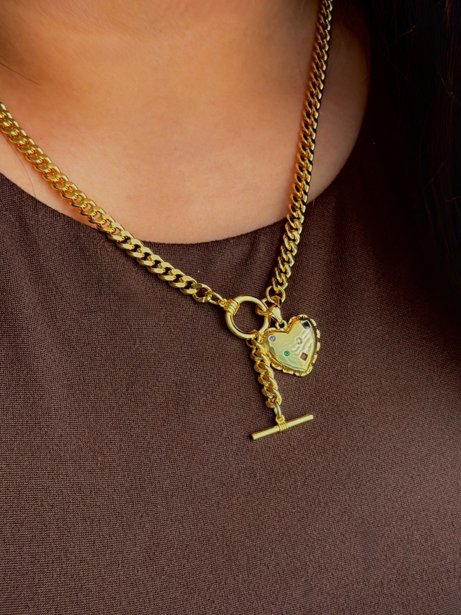 Close-up of a person wearing two gold necklaces, one with a heart-shaped pendant and the other with a T-shaped pendant, against a dark top.