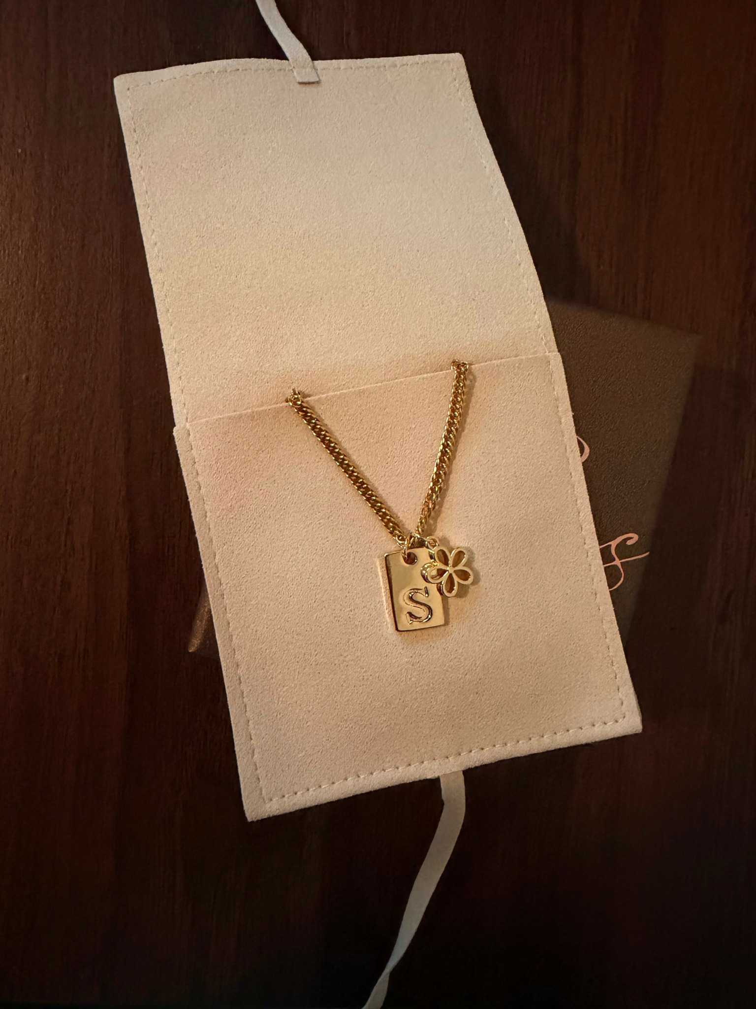 Necklace with a square pendant featuring a flower and letter 'S' inside a jewelry box, open on a wooden surface.