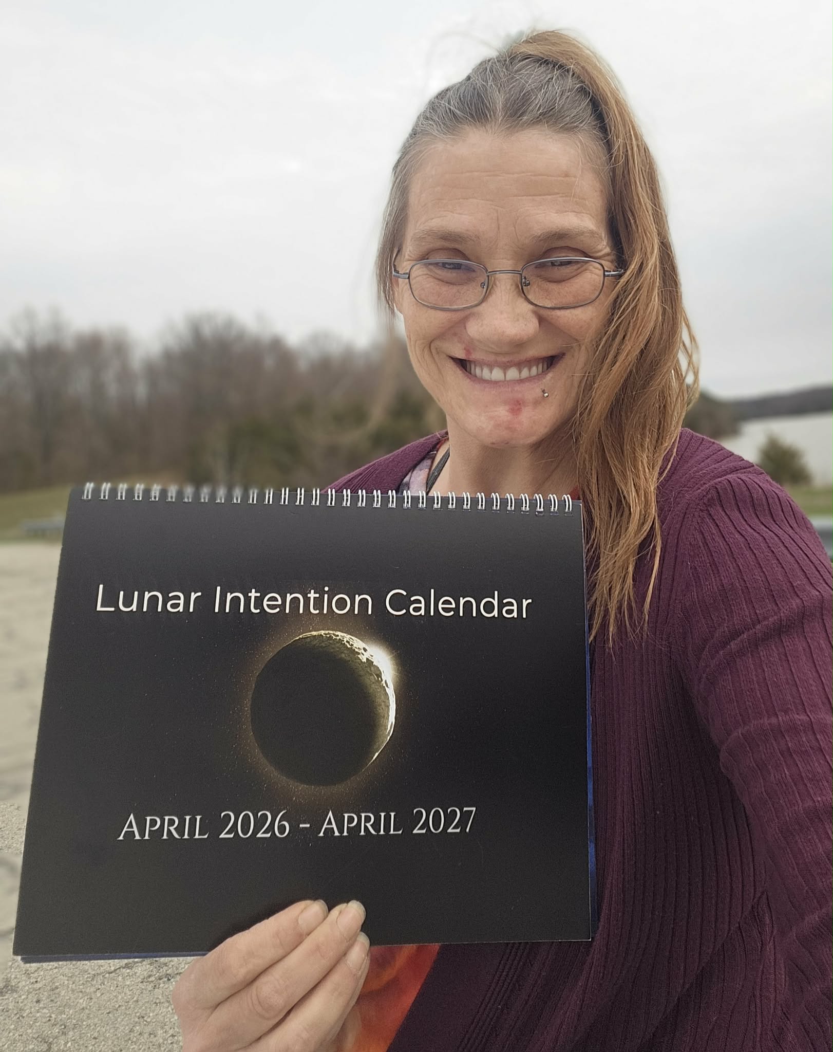 Woman outdoors holding a lunar calendar for April 2026 to April 2027, smiling, with trees and water in background.