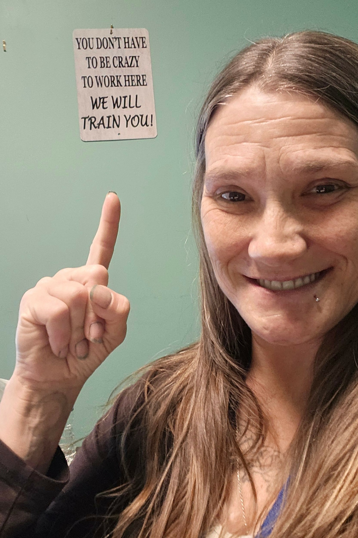 Woman smiling, pointing upward with her index finger, green wall background, motivational sign, long hair, casual clothing.