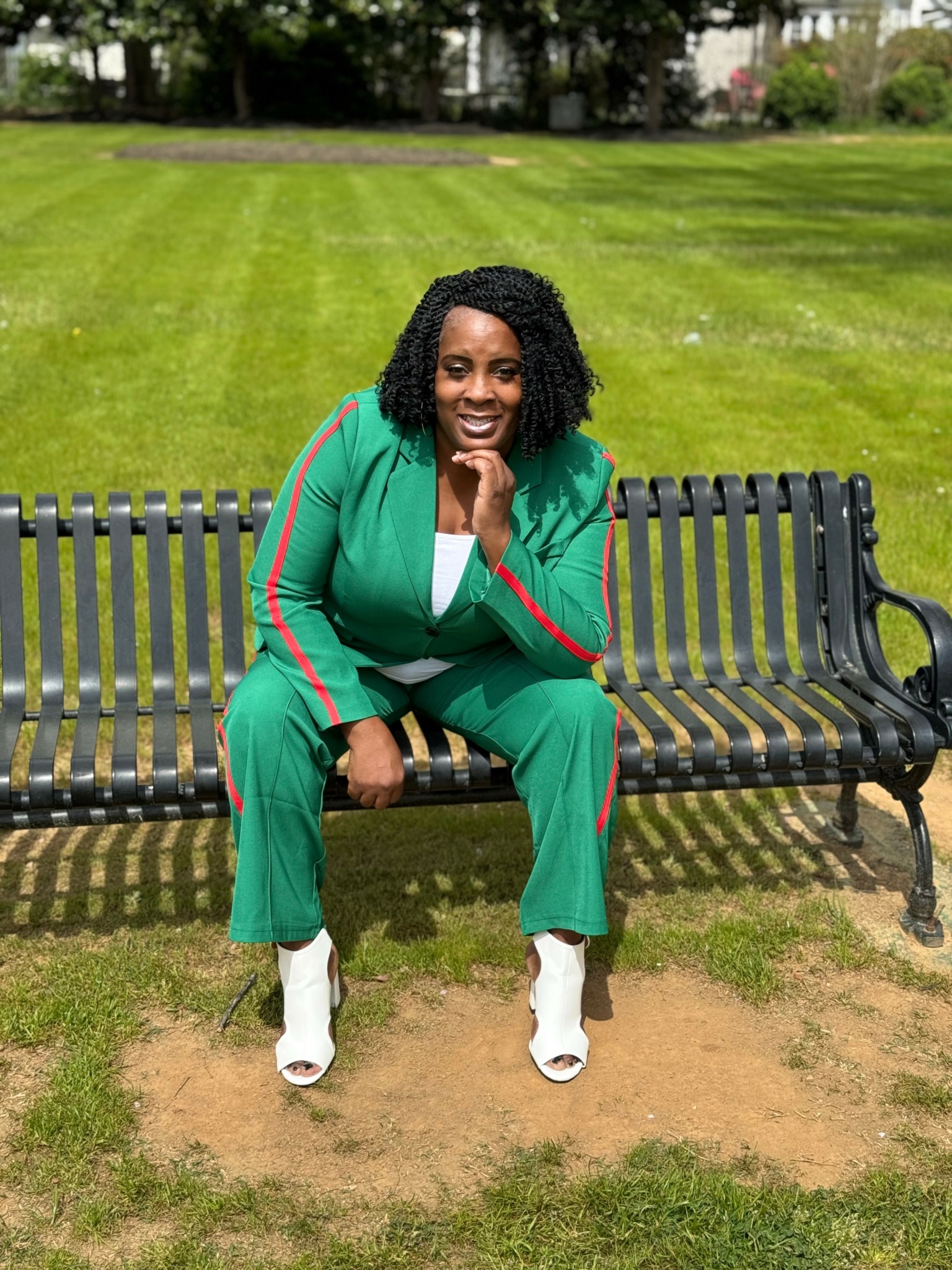 Woman in green outfit sitting on a park bench smiling, with grass and trees in the background.