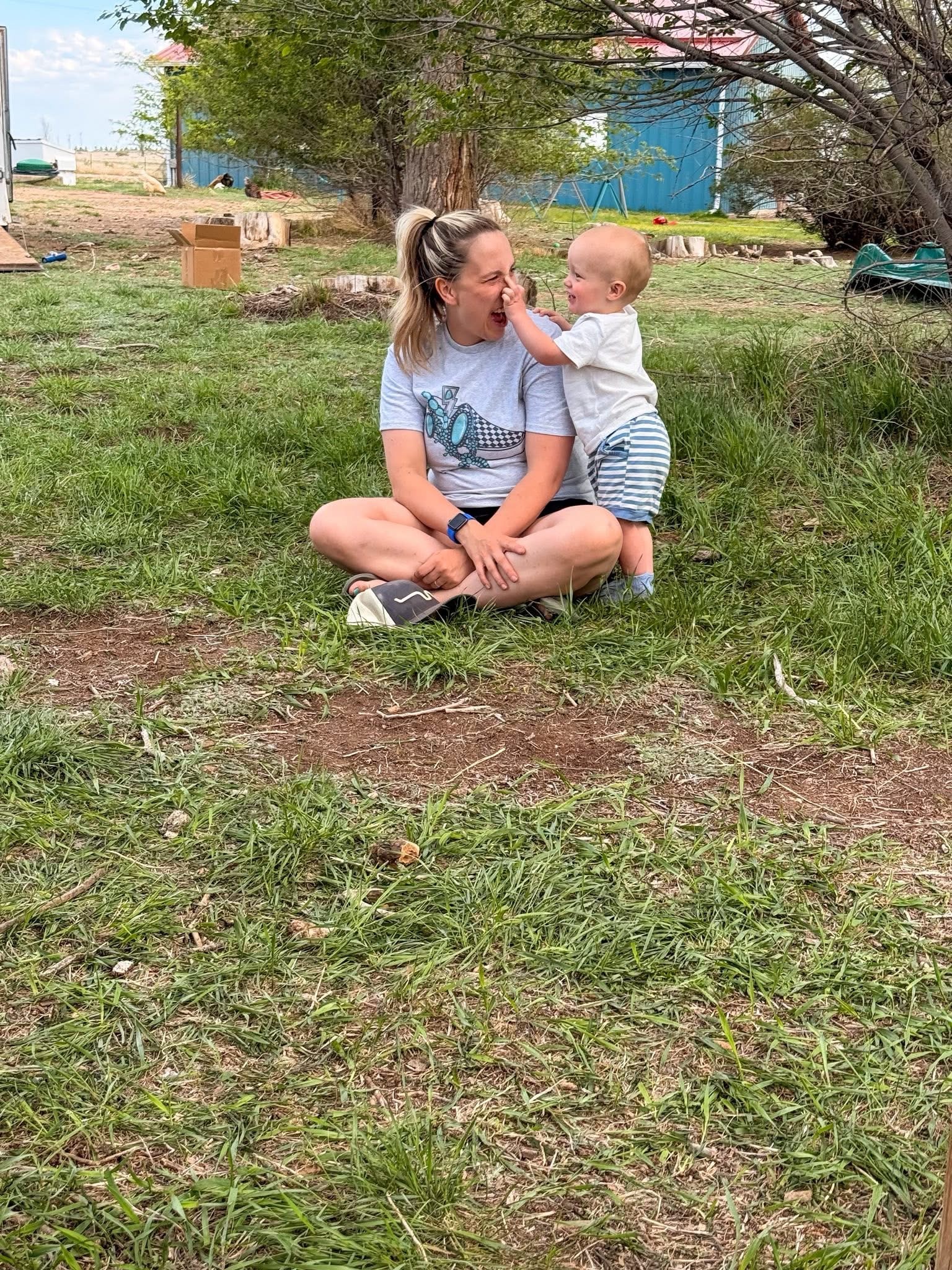 Woman sitting on grass with a young child, trees and a blue building in background, outdoors during daytime.