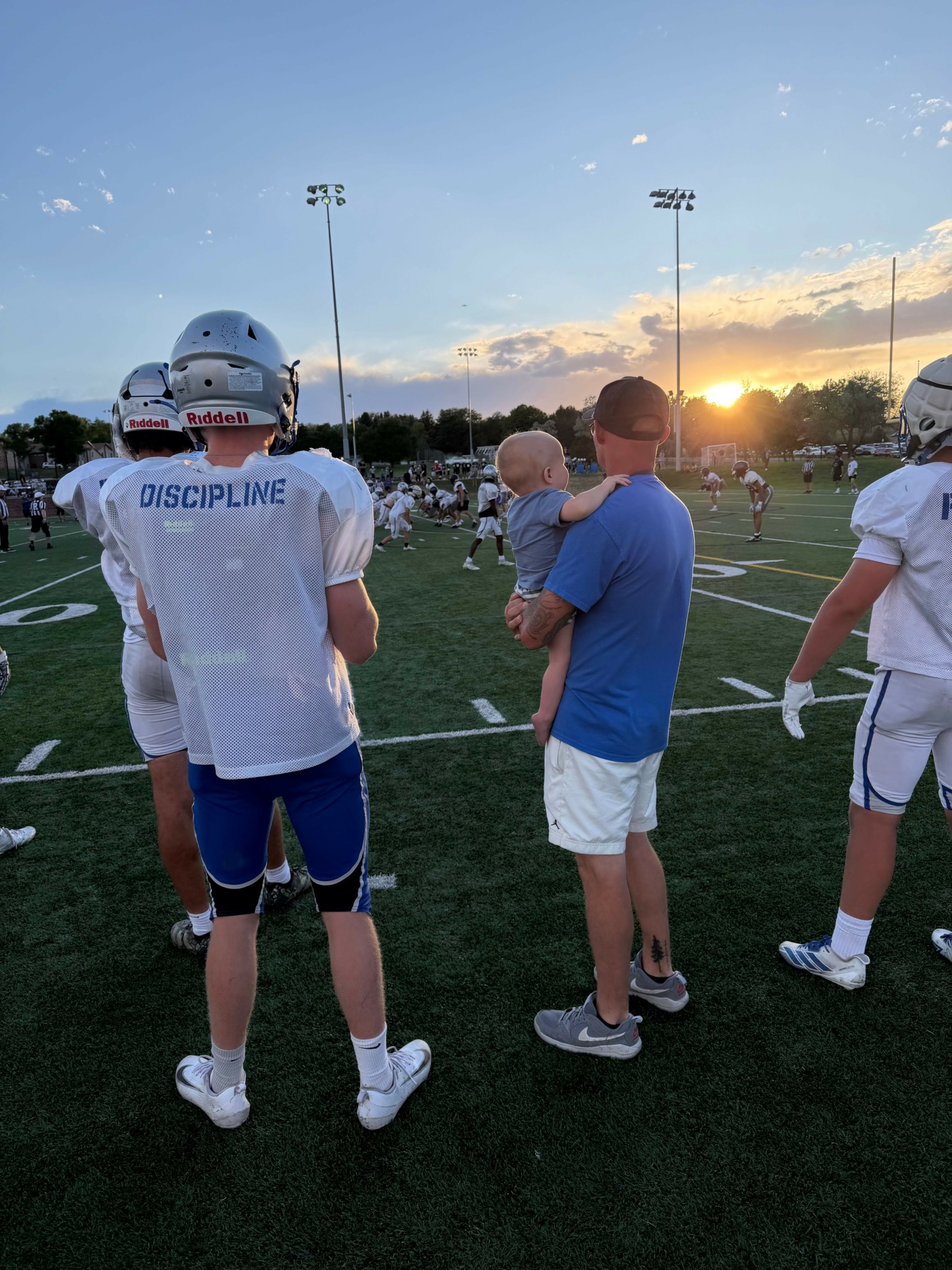 Two football players on a field during sunset, one in a white jersey and the other in a blue shirt, facing each other.