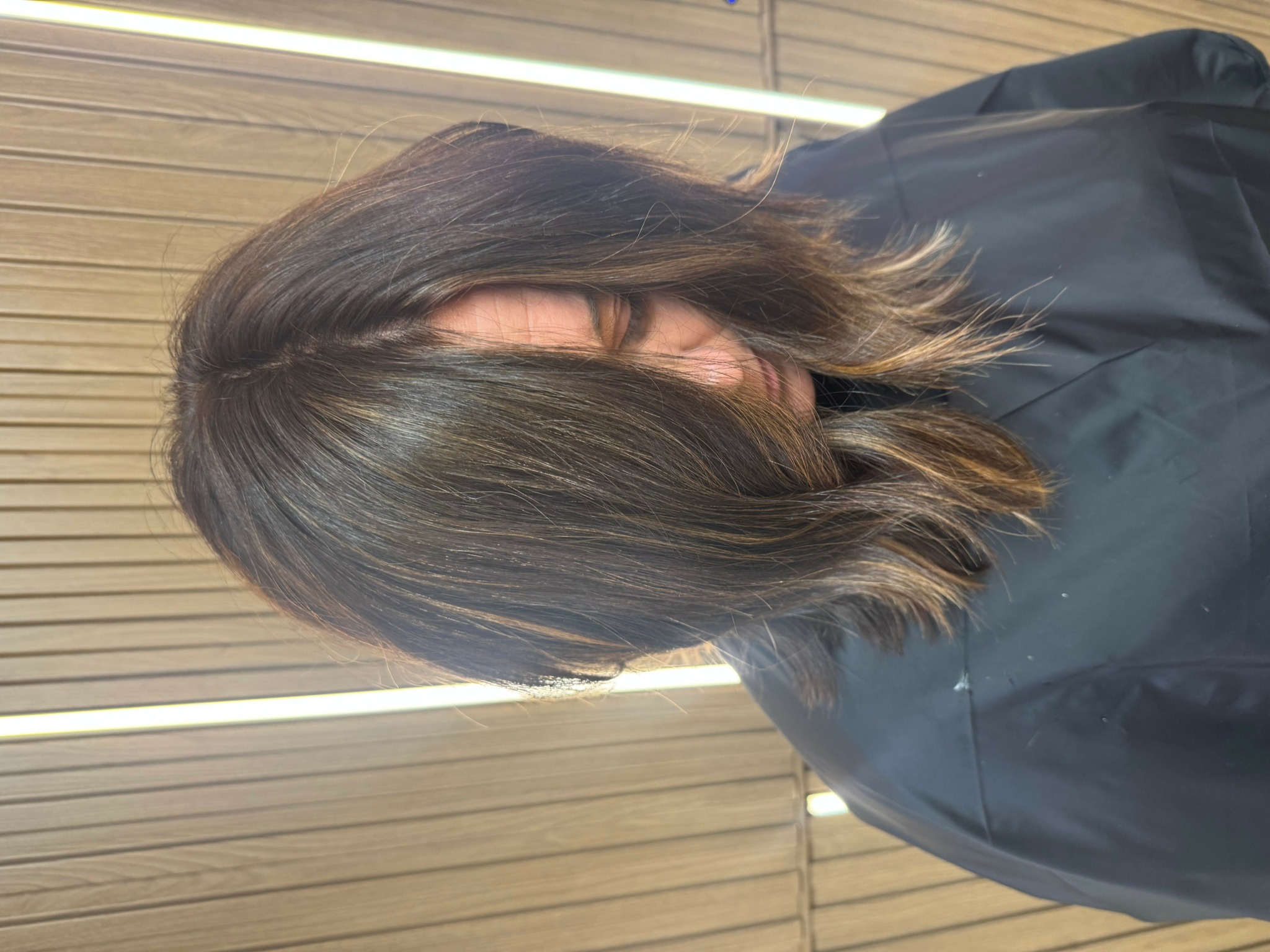 Person with shoulder-length dark hair, wearing a black jacket, standing in front of wooden panels.