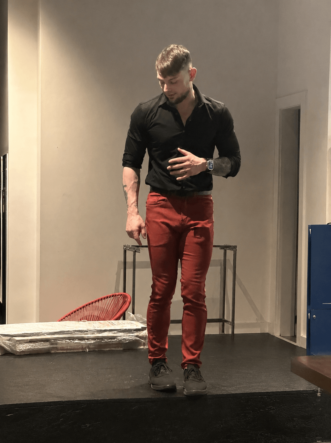 Young man standing on stage, looking down, wearing black shirt and red pants, with a plain background.