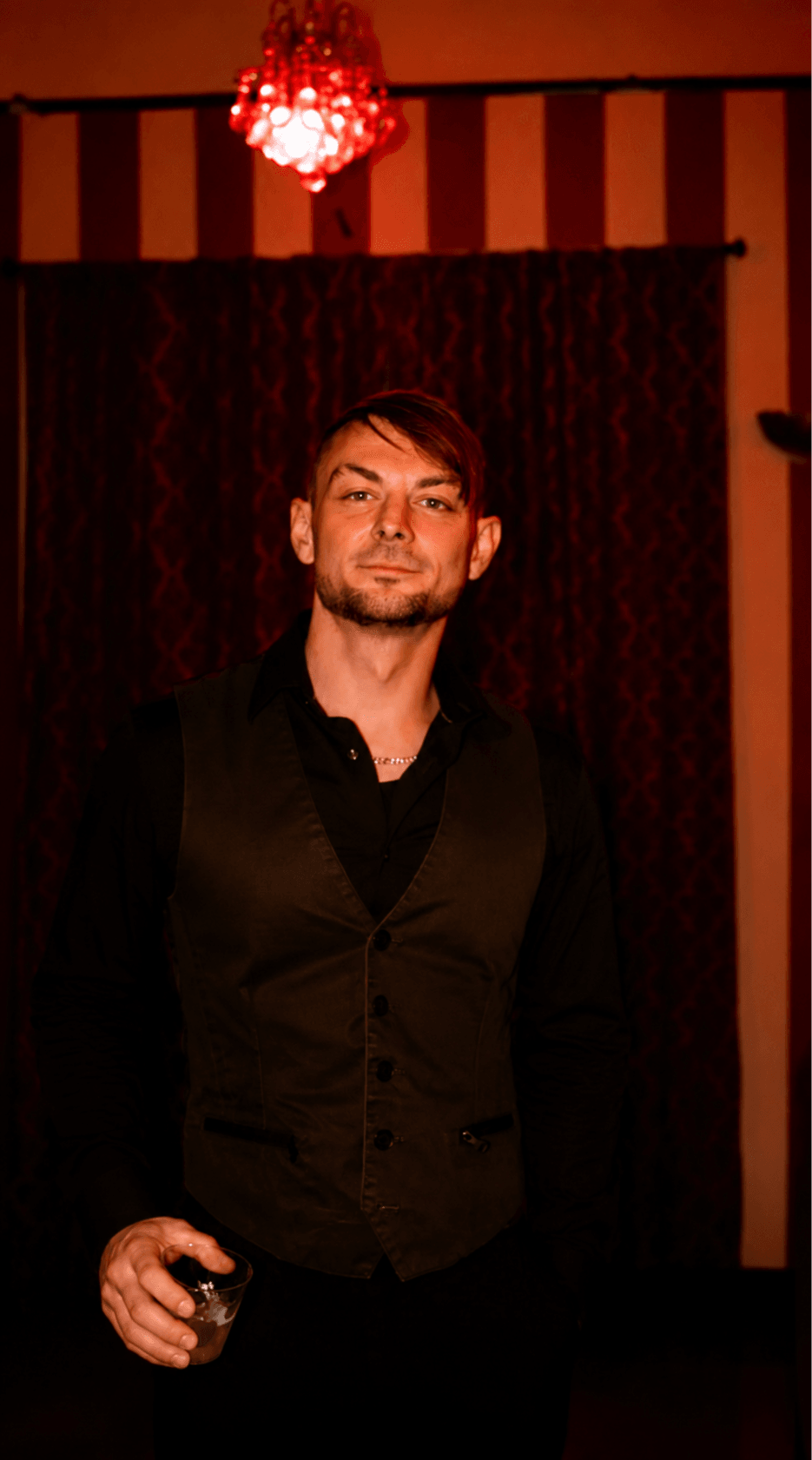Man with short hair and beard in a dark vest and shirt, standing indoors with striped wallpaper and a red chandelier.
