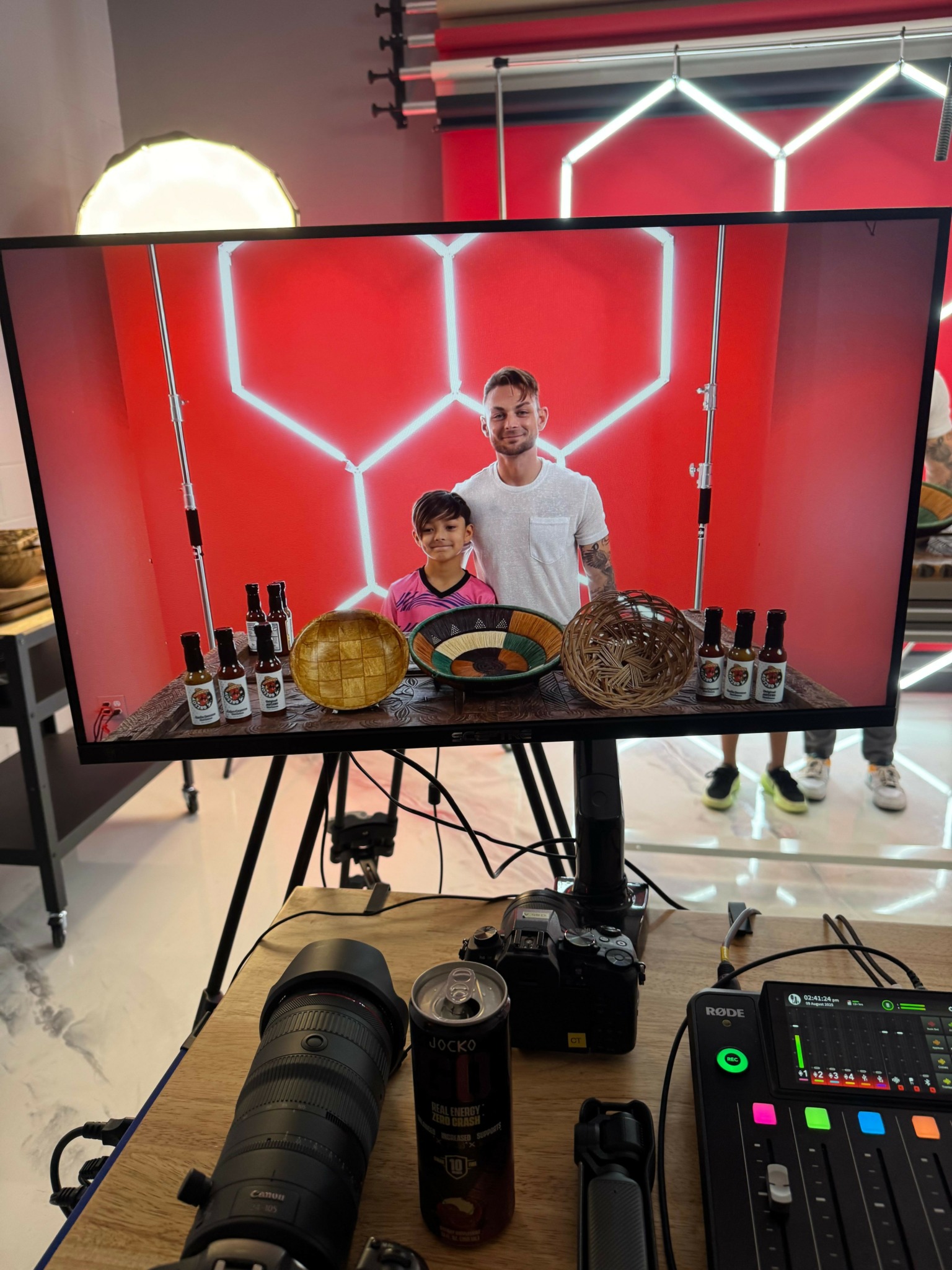 Two people stand behind a table with drinks and snacks in front of a red hexagonal patterned background.