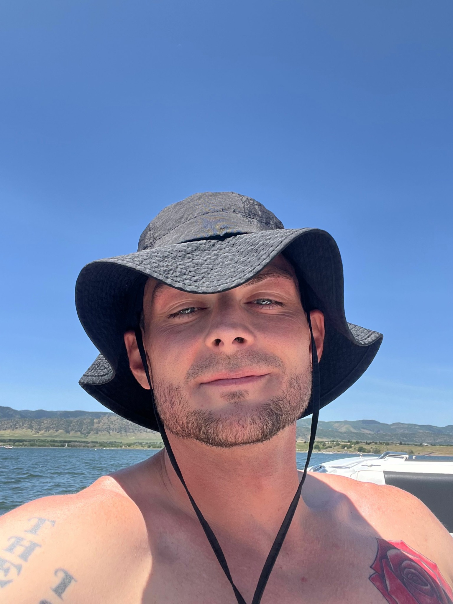 Man wearing a wide-brimmed hat and gray cap, smiling at the camera, with a beach and ocean in the background.
