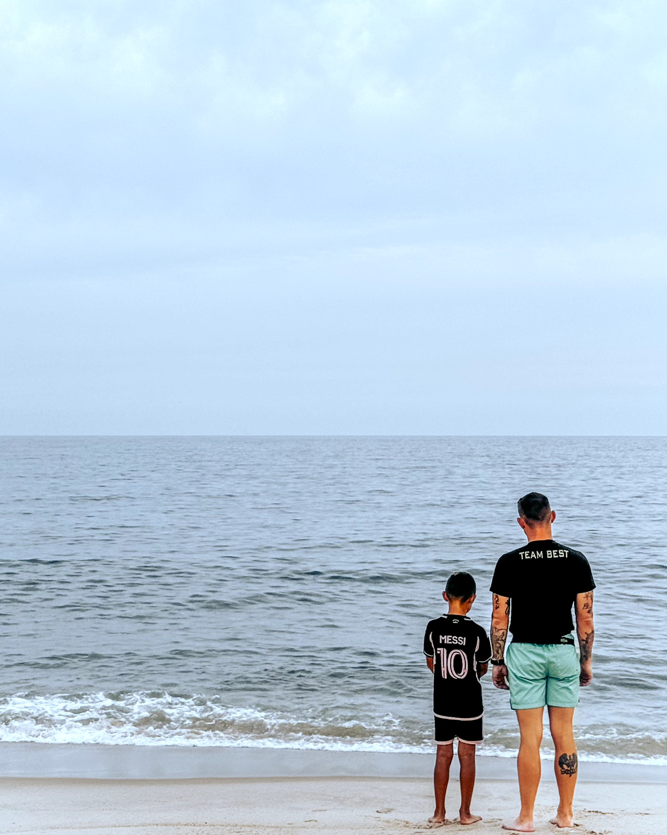 Two people, an adult and a child, stand on the beach facing the ocean under a cloudy sky.