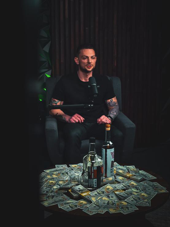 Man with tattoos sitting in a dark room, surrounded by money and alcohol bottles, speaking into a microphone.