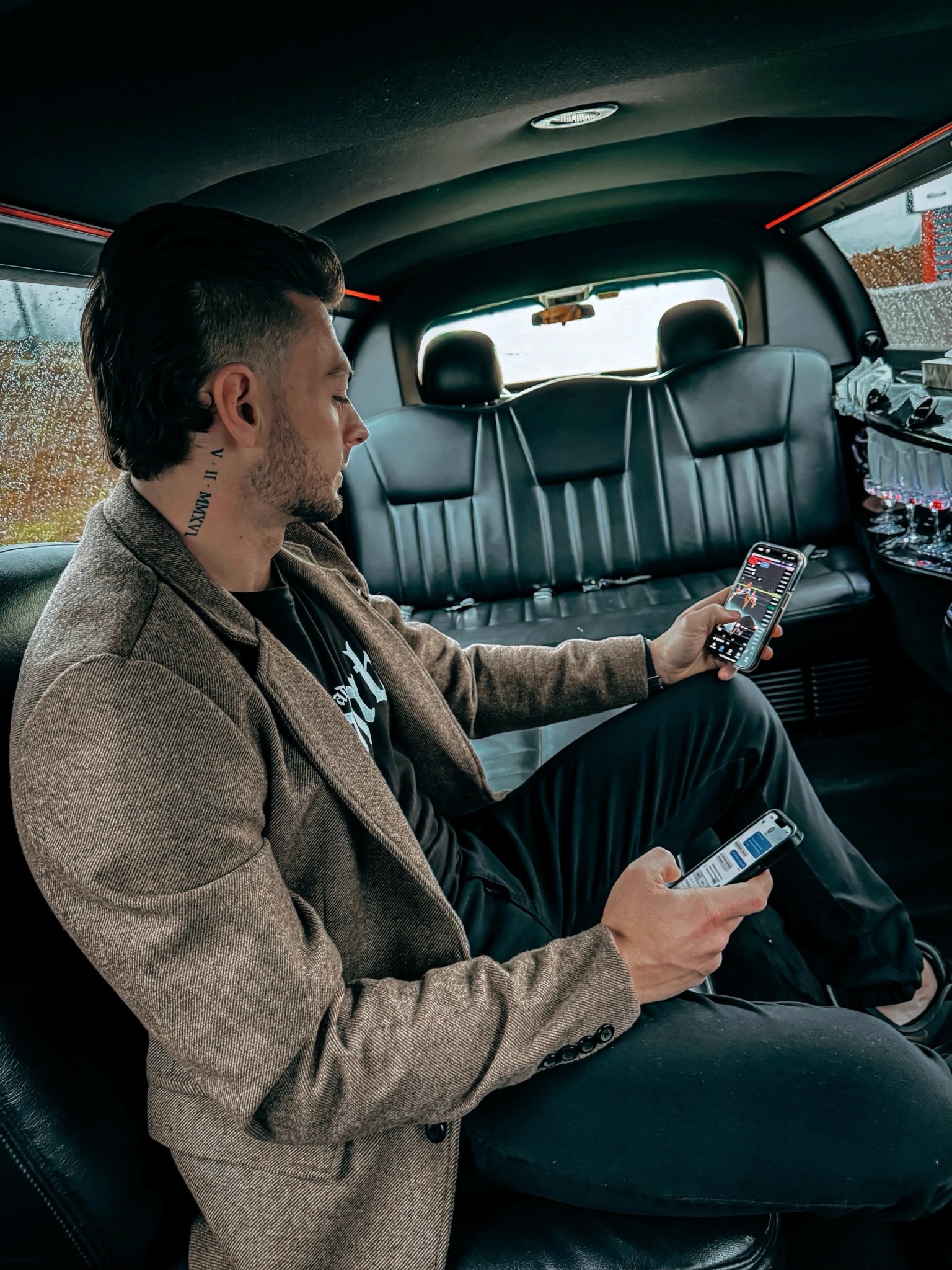 Man sitting in car backseat, looking at phone, with two phones in hands, wearing a brown coat and black pants.
