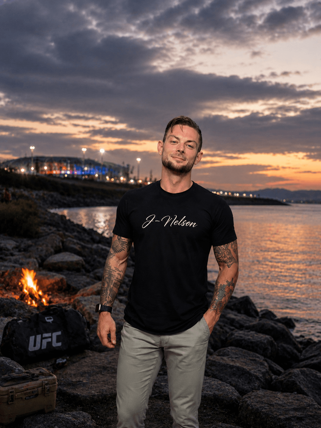 Man standing on rocky shoreline during sunset, wearing a black T-shirt and light-colored pants, with tattoos on arms.