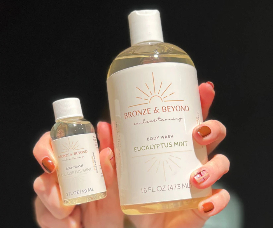 Person holding two bottles of body wash, one large and one small, with black background. Both bottles are labeled 'Bronze & Beyond' and 'Eucalyptus Mint'.