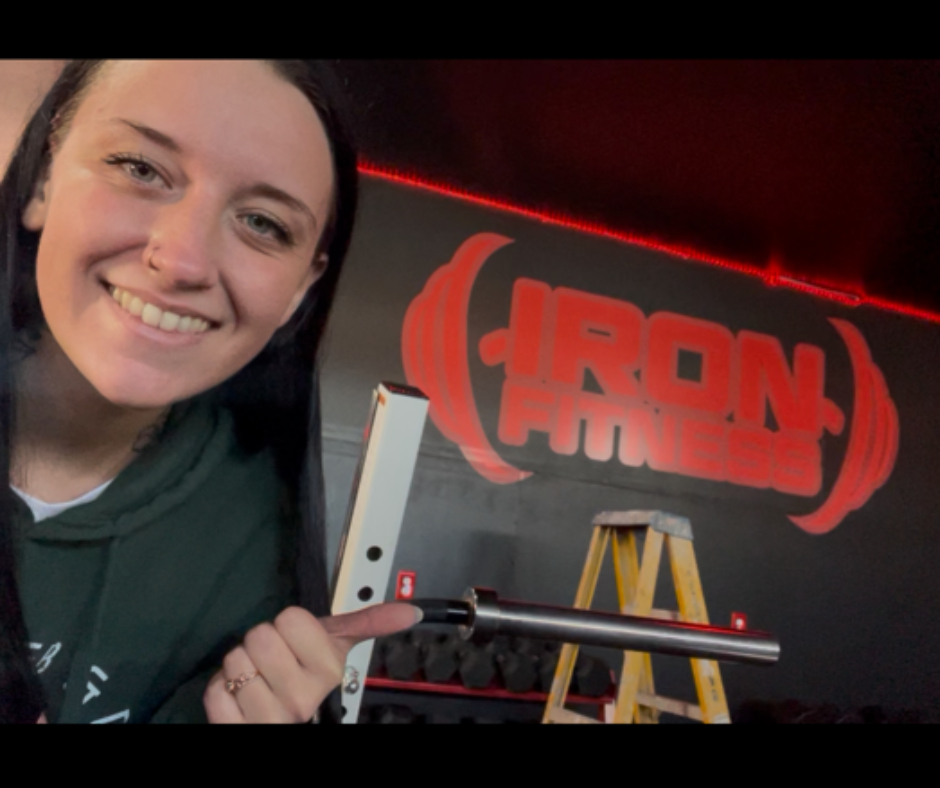 Smiling woman holding a long object in front of a sign that says 'IRON' with a red background.