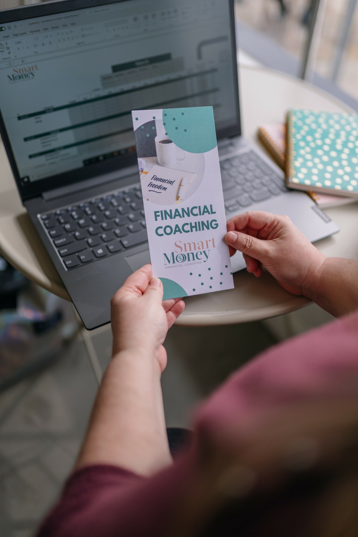 Person holding a financial coaching brochure with a laptop and notebook on a table.