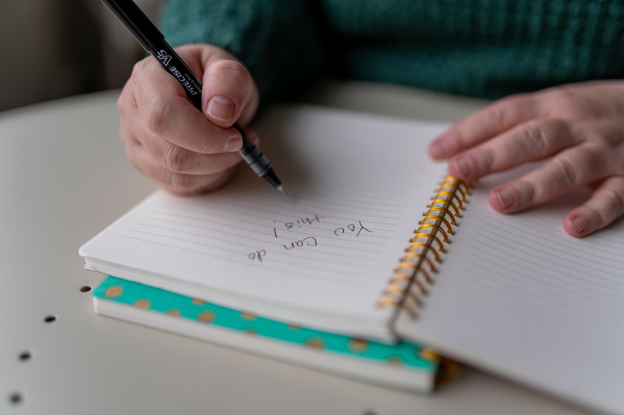Person writing in a notebook with a pen, resting hands on the table, notebook open, lined pages visible.