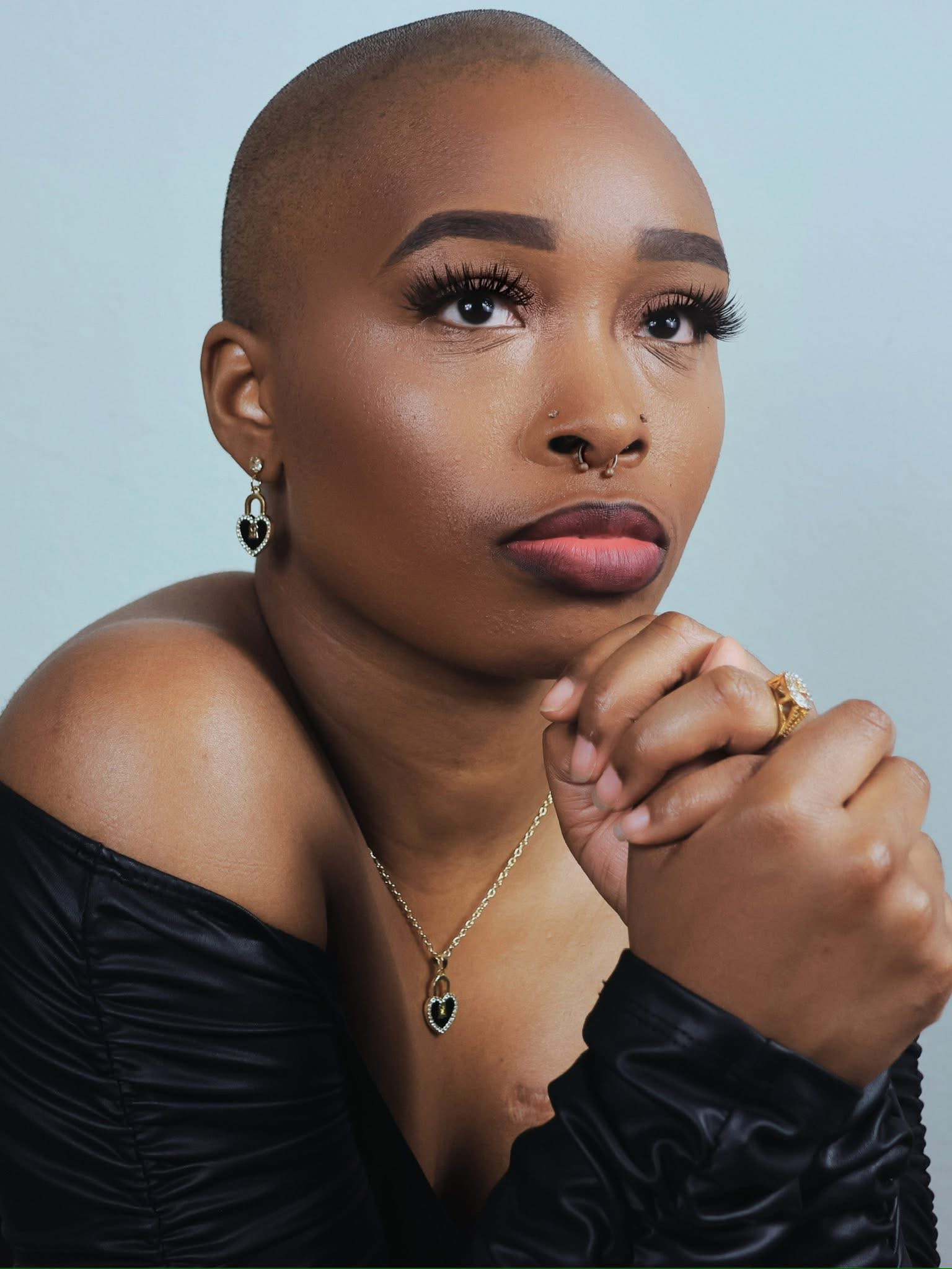 Portrait of a woman with a shaved head, wearing earrings, a nose ring, and a necklace, looking thoughtfully with hands clasped.