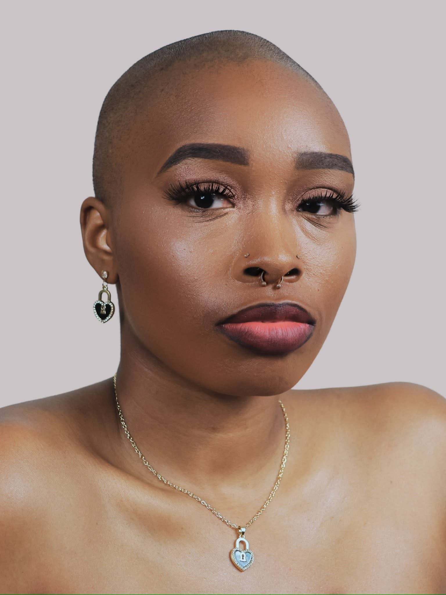 Close-up of a person with a shaved head, makeup, earrings, and a necklace, looking directly at the camera.
