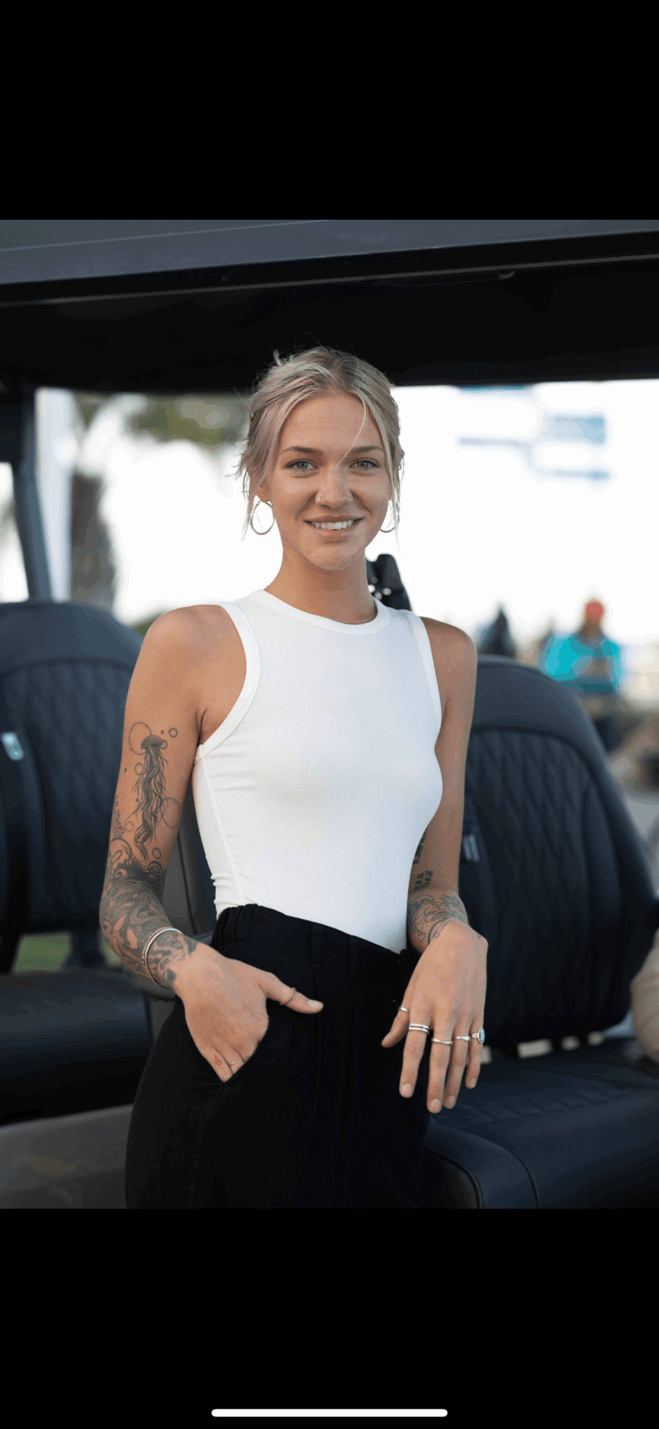 Woman with tattoos on arms, smiling, wearing a white sleeveless top, standing outdoors near a vehicle.