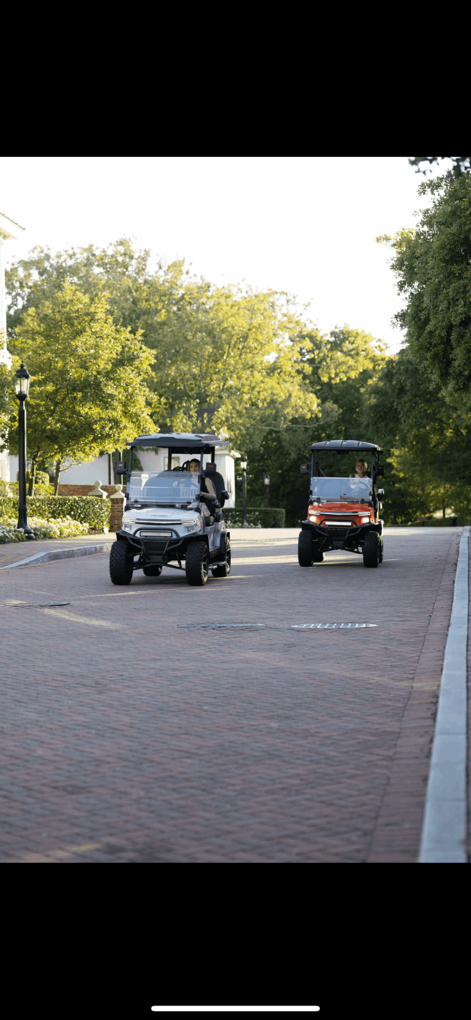 Two golf carts parked on a paved area with trees and a lamppost in the background.