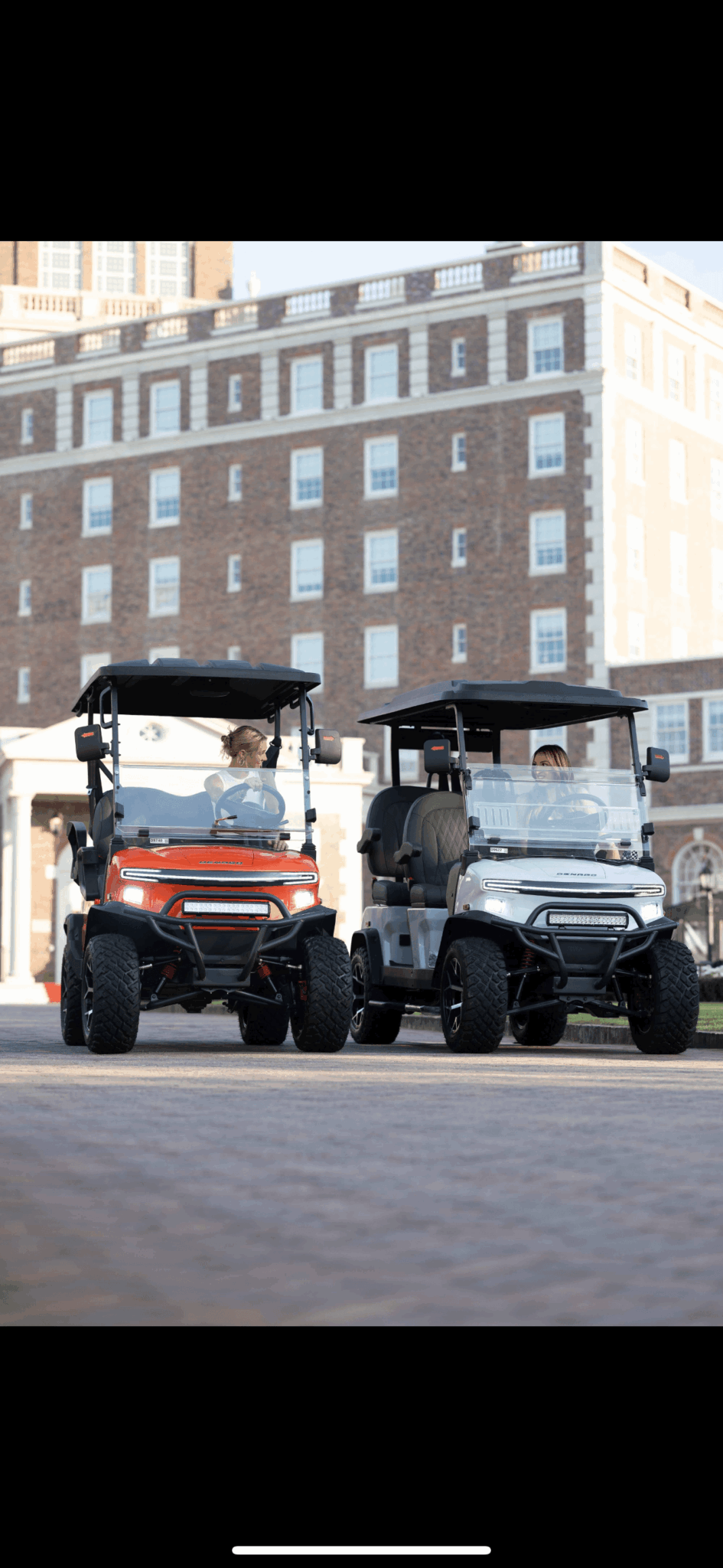 Two golf carts parked side by side in front of a large building with multiple windows and brick facade.