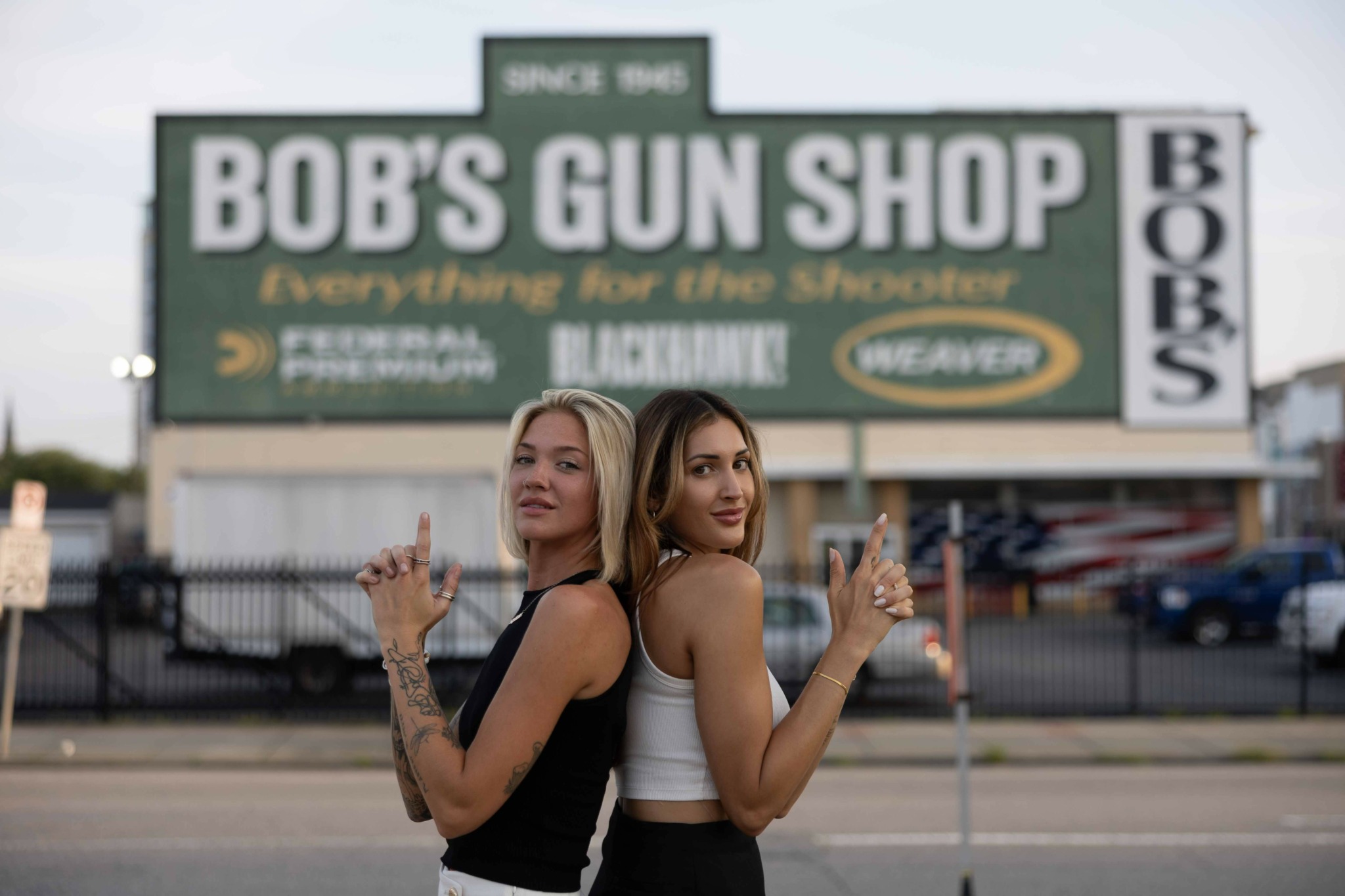 Two women stand back-to-back in front of a gun shop sign, holding guns and looking to the side.