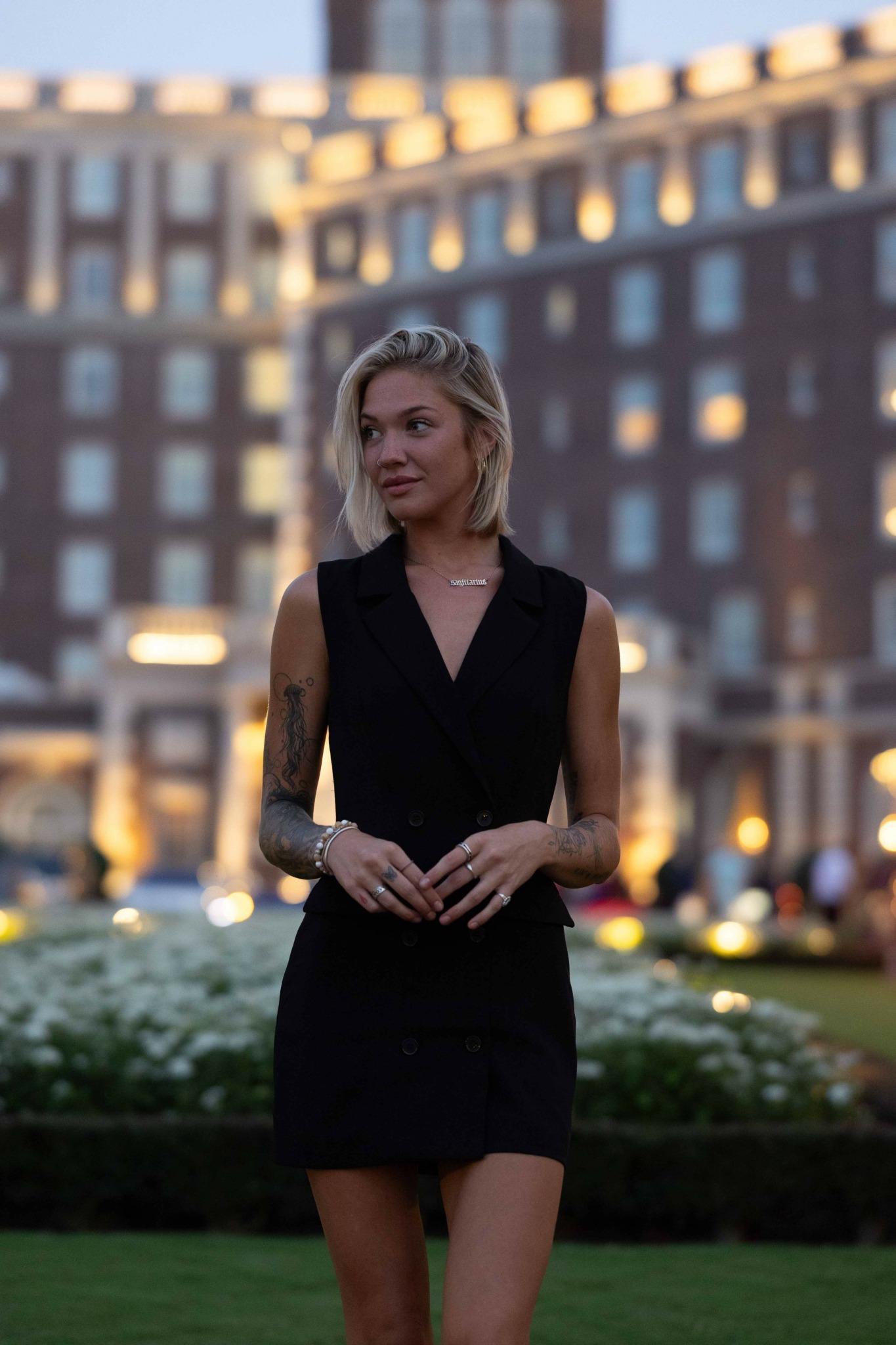 Woman in black dress standing outdoors with buildings in background, during evening or dusk.