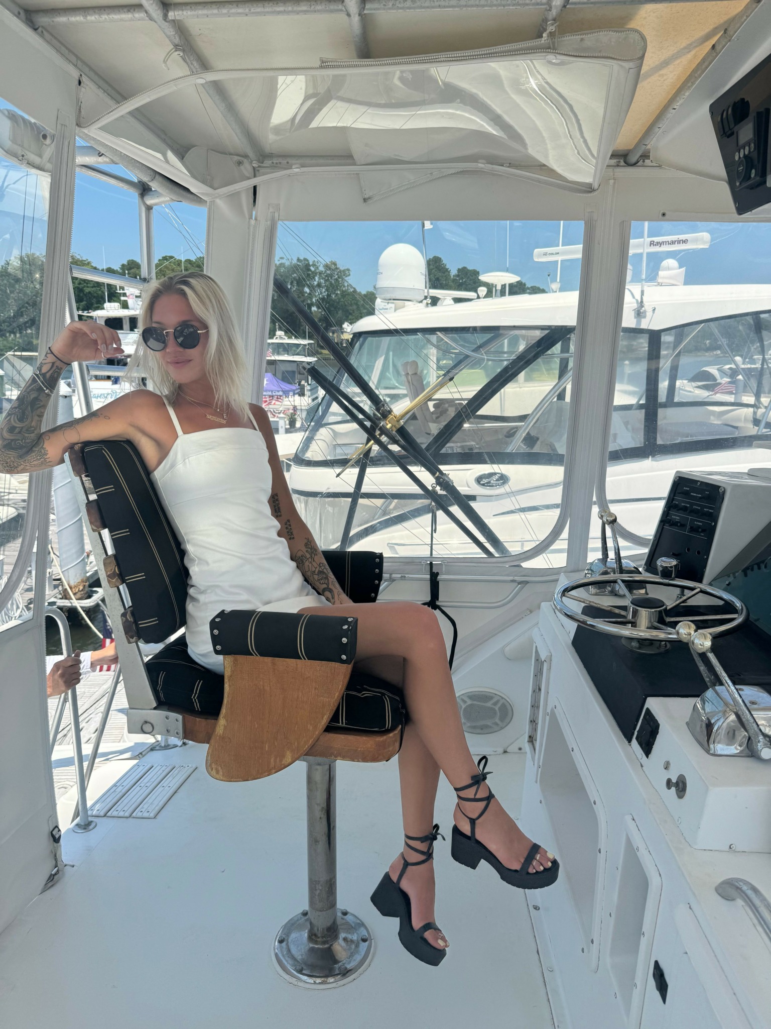 Woman with blonde hair in a white dress sitting on a chair in a boat's control room, holding a drink, with a view of water and boats outside the window.