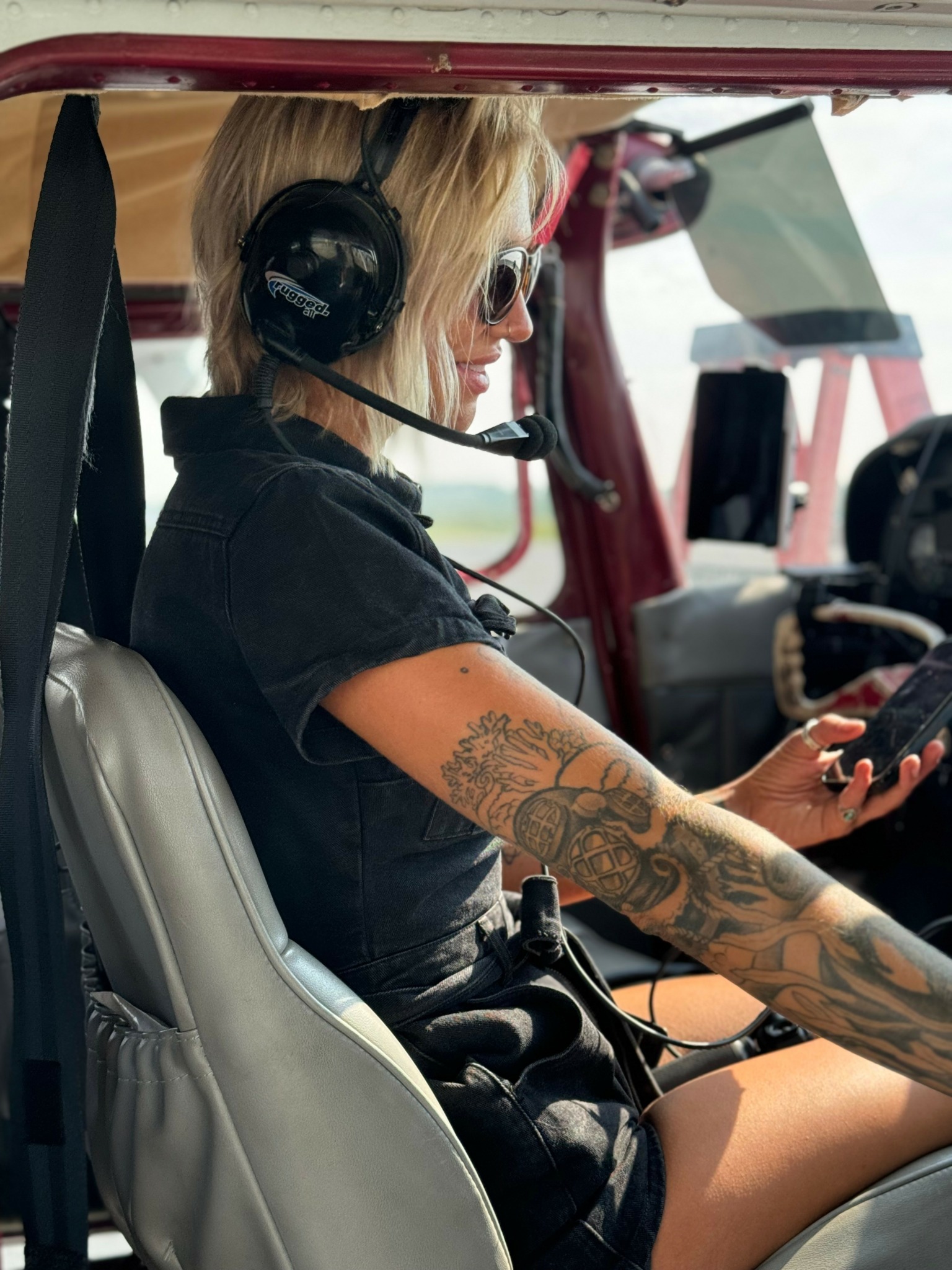Woman with tattoos wearing headphones, sitting in vehicle, looking at phone, with a red vehicle interior visible.
