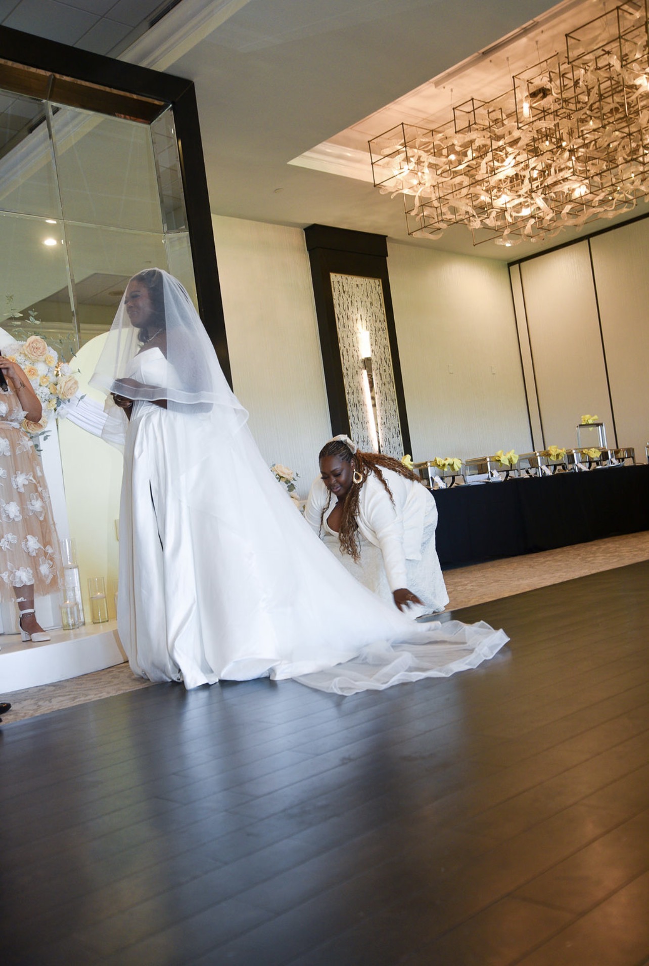 Bride in a white wedding gown and veil stands with a person in a white dress, in a decorated indoor venue.