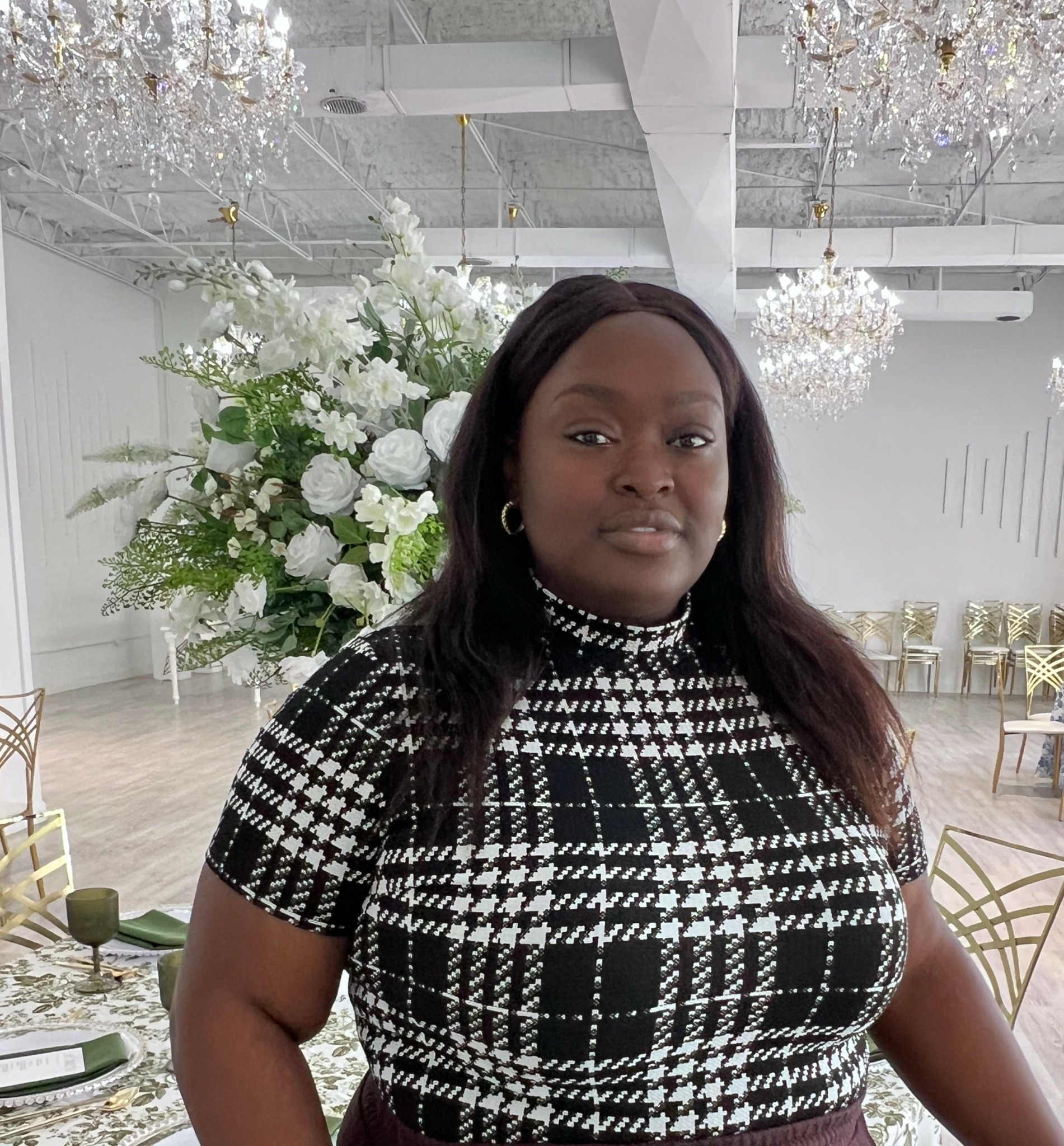 Woman with dark hair in a black and white patterned top standing in a decorated event space with floral arrangements and chandeliers.