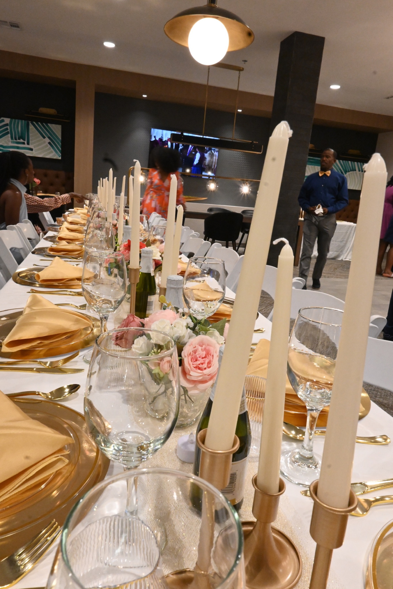 Elegant table setting with candles, flowers, and glassware in a decorated dining area with people in the background.