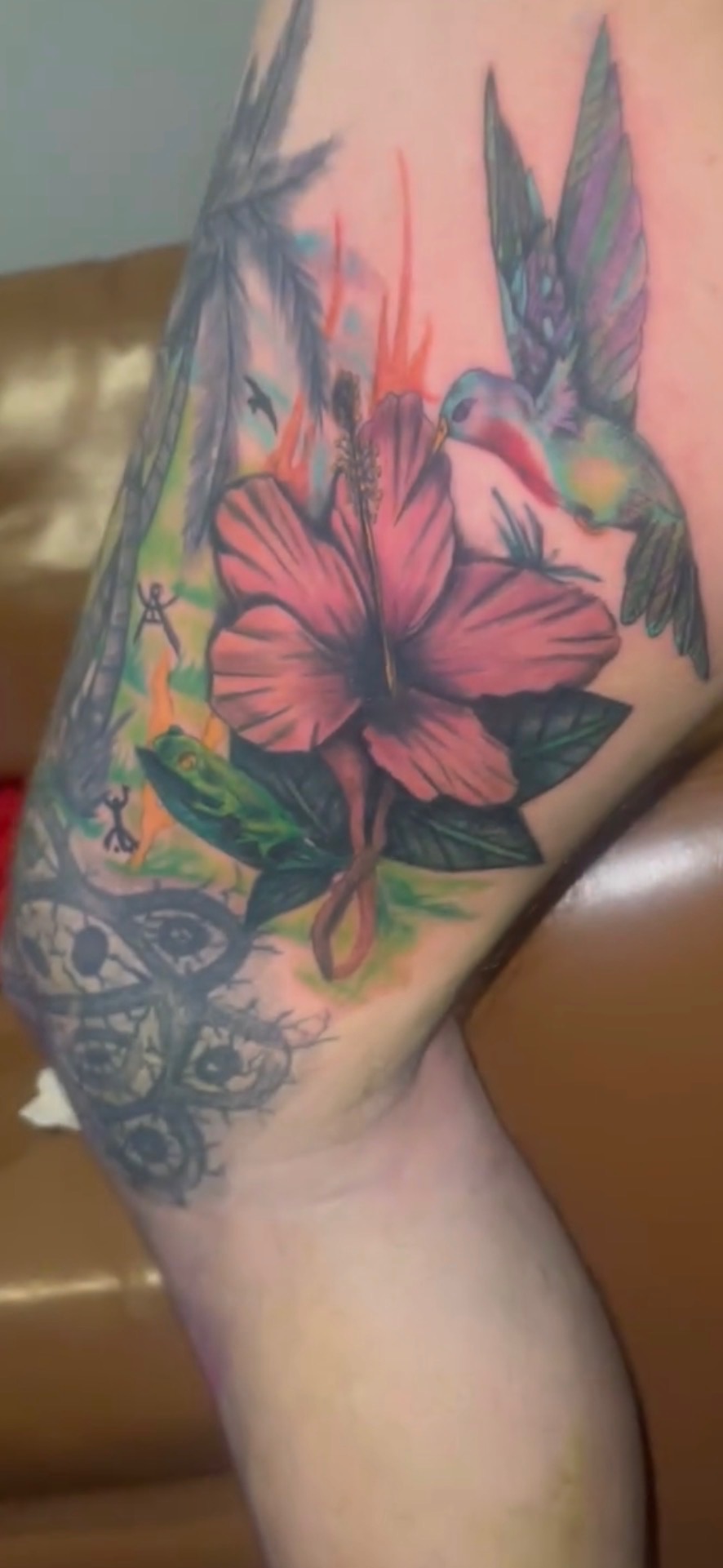 Tattoo of a hummingbird, pink flower, and green leaves on a person's arm, with colorful background elements.