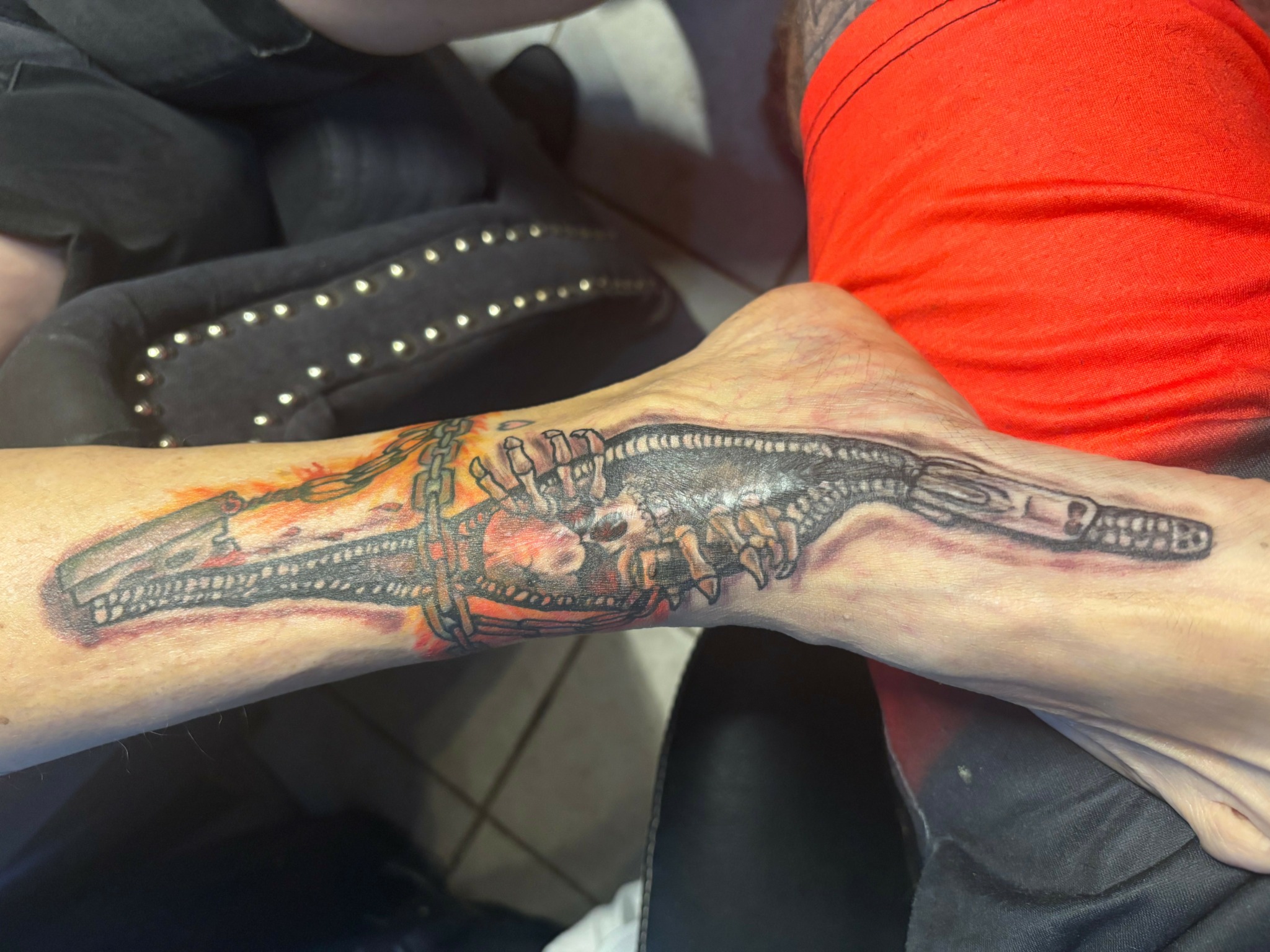 Tattooed arm with a detailed, colorful design, resting on a person's lap, with a black studded bag nearby.