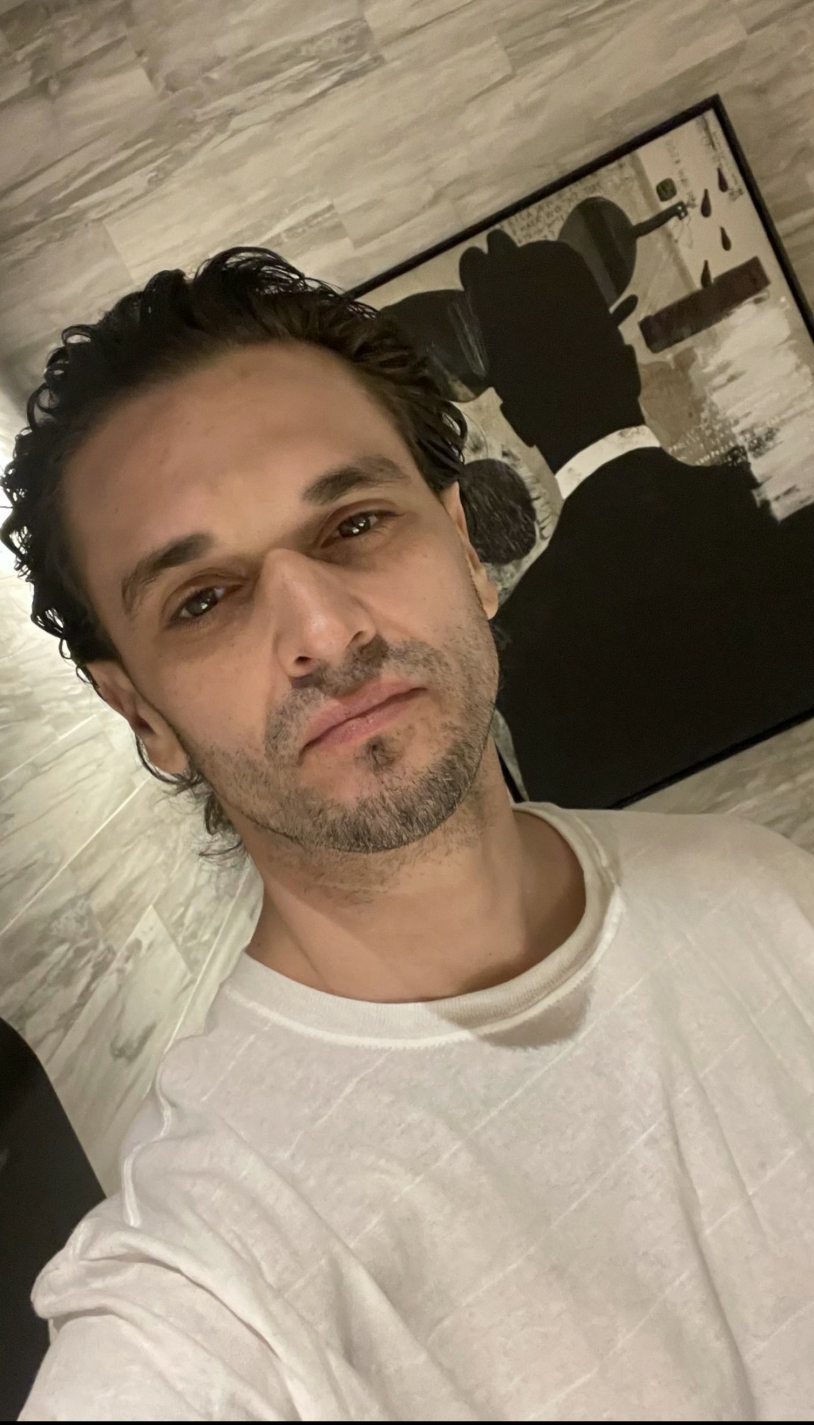 Man with dark curly hair and light beard taking selfie indoors, with a black and white silhouette artwork in the background.